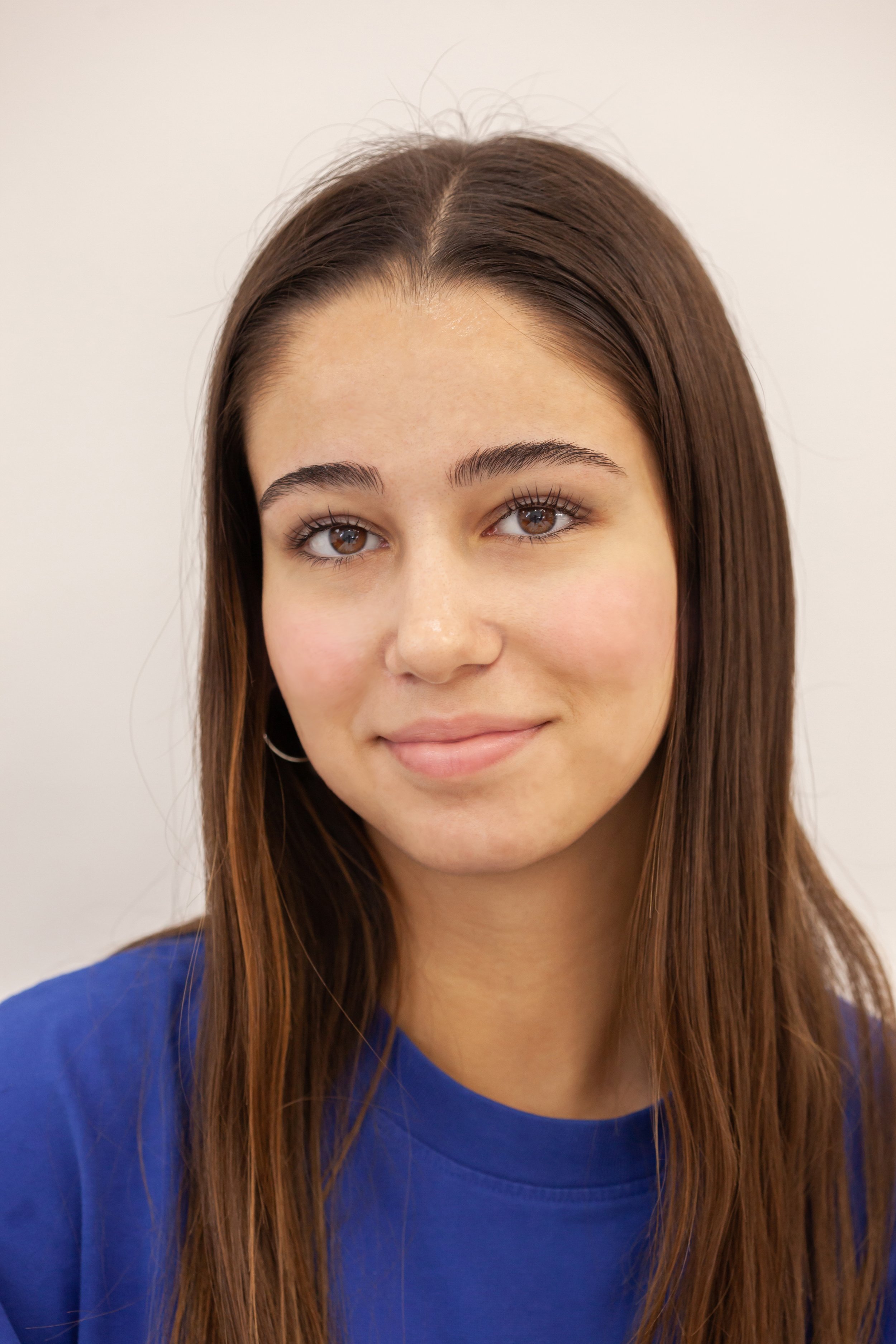 A young woman with long brown hair, wearing a blue shirt, smiling softly with a light skin tone, brown eyes, and small hoop earrings, standing against a plain off-white background. Dbeaute studio. Lash & Brow Lift. Powder brows. Makeup art. Dbeaute s