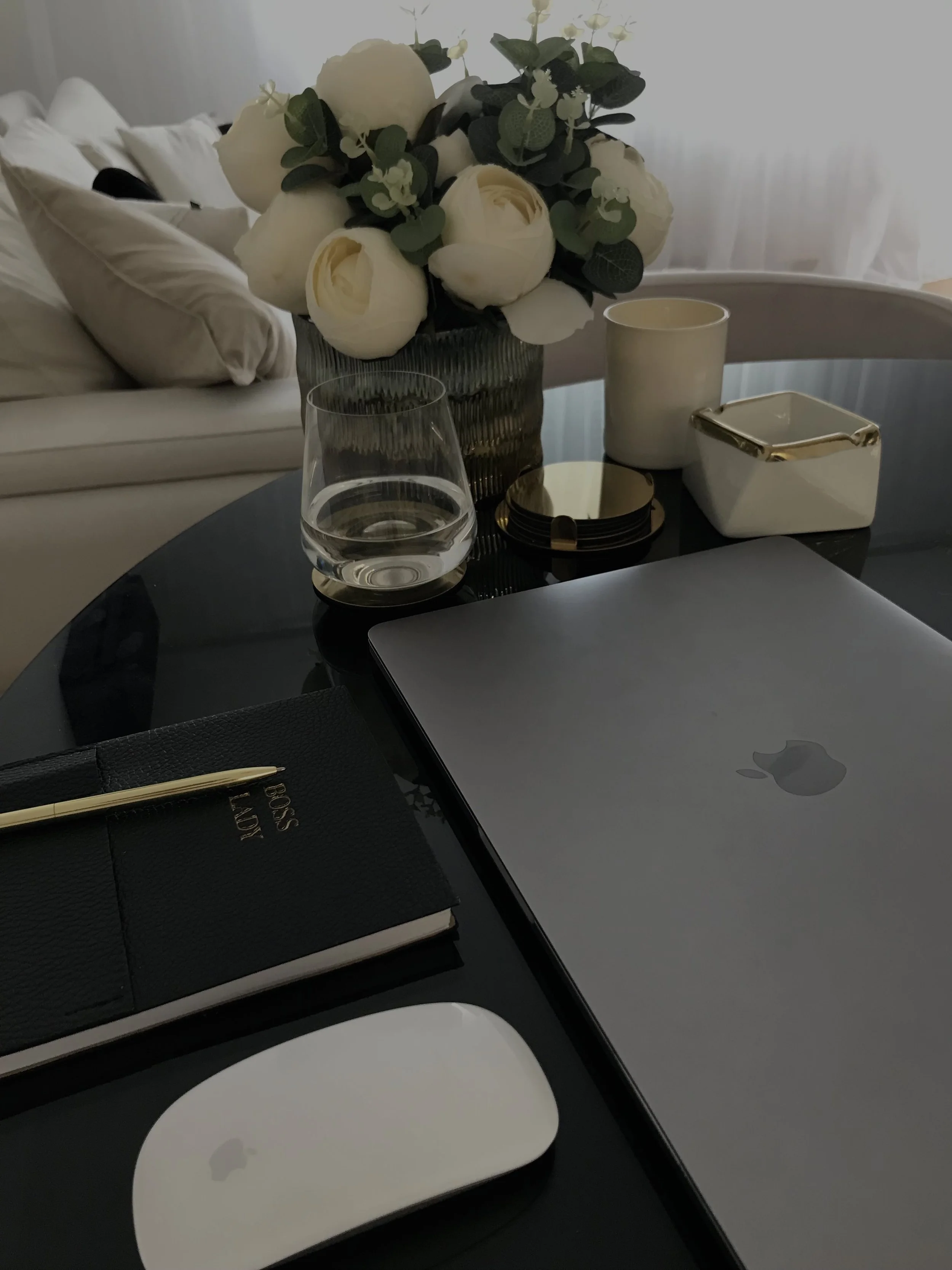 Desk with closed MacBook, Magic Mouse, planner with pen, glass of water, decorative flowers in a vase, white candle, small white and gold box, phone stand, and black coasters.