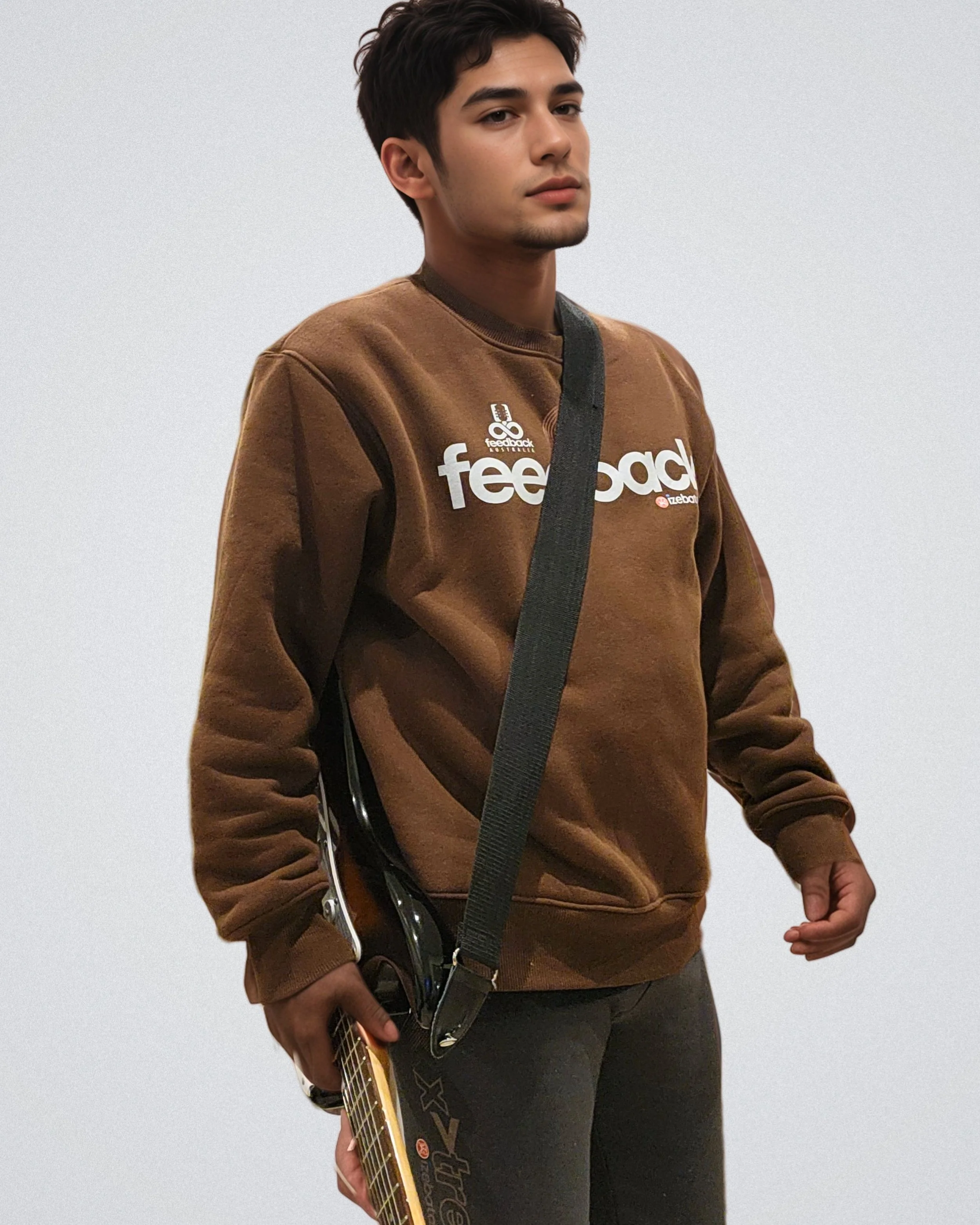 Young man wearing a brown sweatshirt with the word feedback and graphics, holding a guitar with a strap over his shoulder, standing against a plain background.