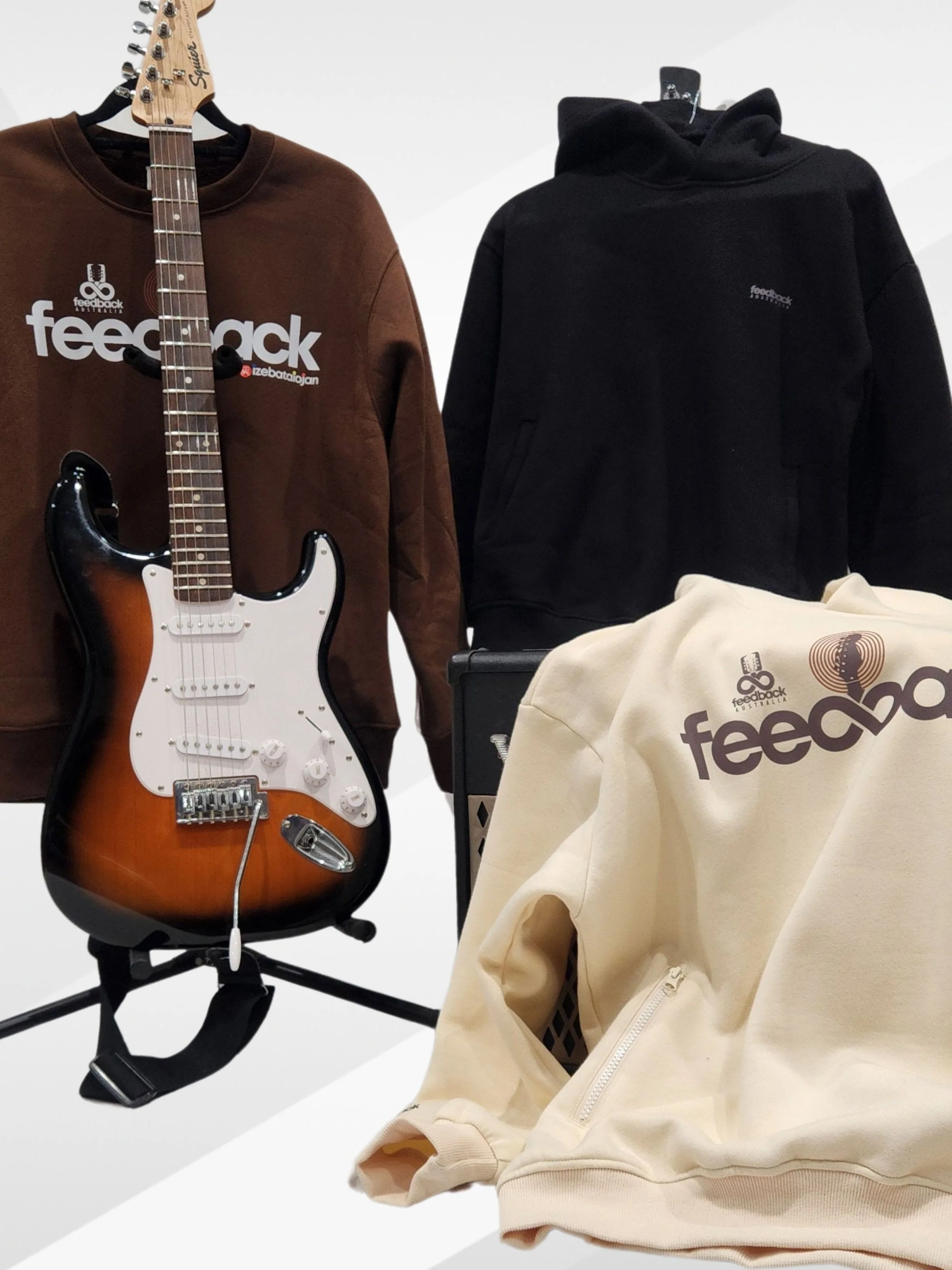 Display of three sweatshirts and a guitar. The brown sweatshirt has 'feedback' written on it with a logo above. The black sweatshirt has a small logo on the chest. The cream-colored sweatshirt has 'feedback' written with a logo above. An electric gui