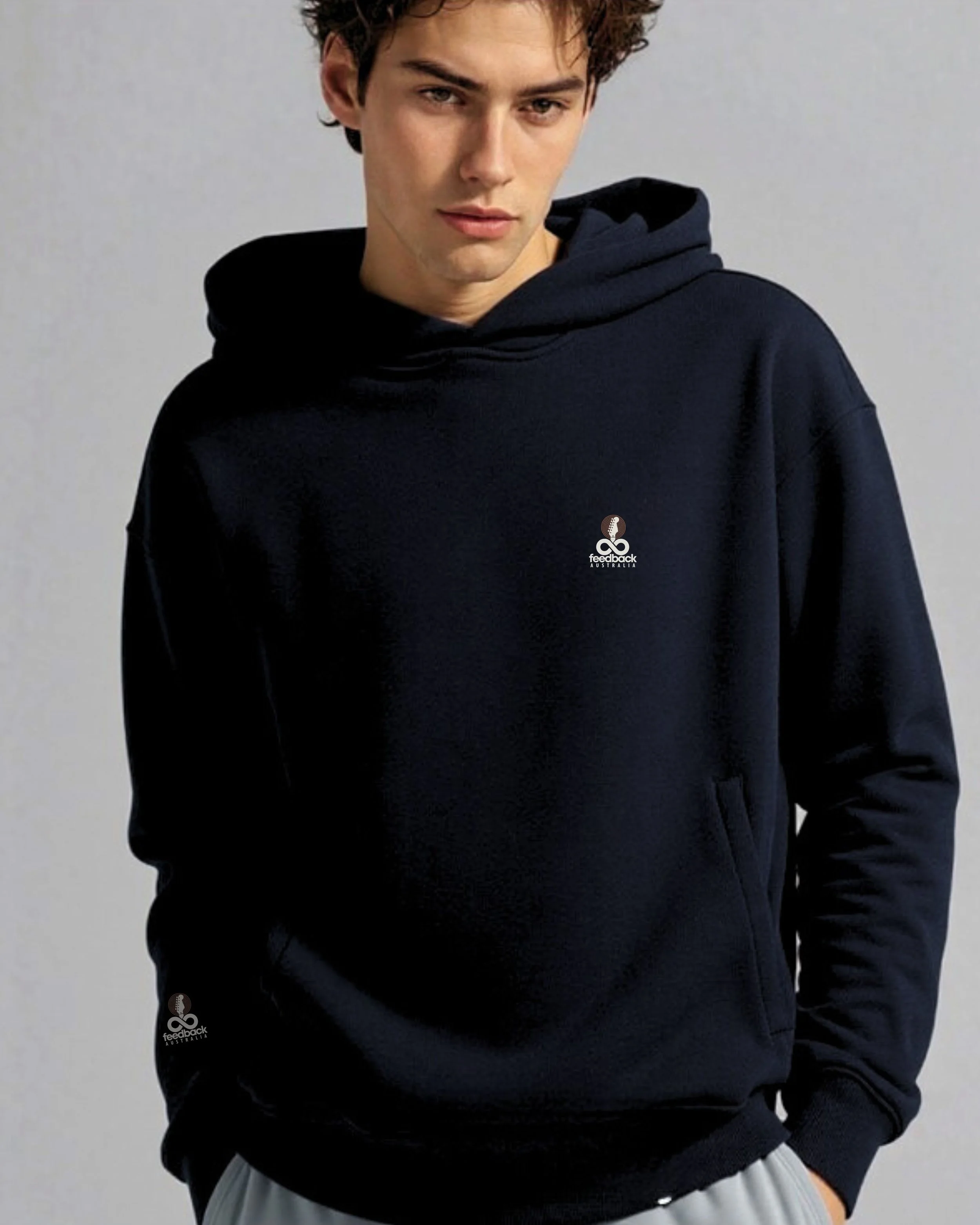 A young man with curly brown hair wearing a black hoodie with a small logo on the chest and sleeve, standing against a plain gray background.