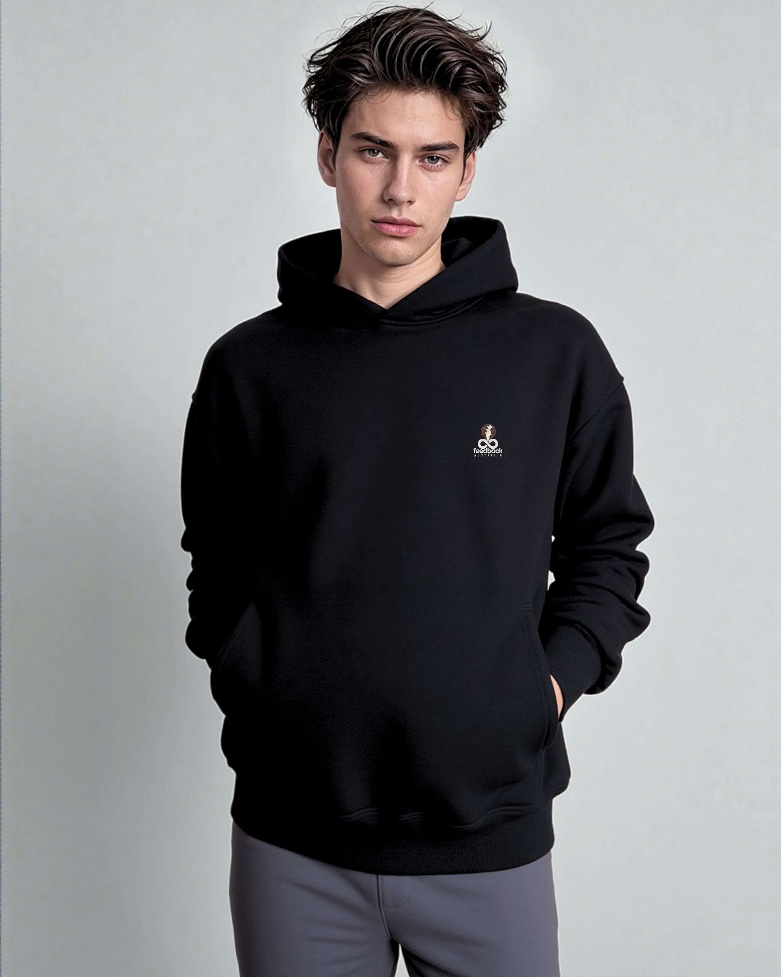 Young man with brown hair wearing a black hoodie with a small logo and grey pants, standing against a plain light background.