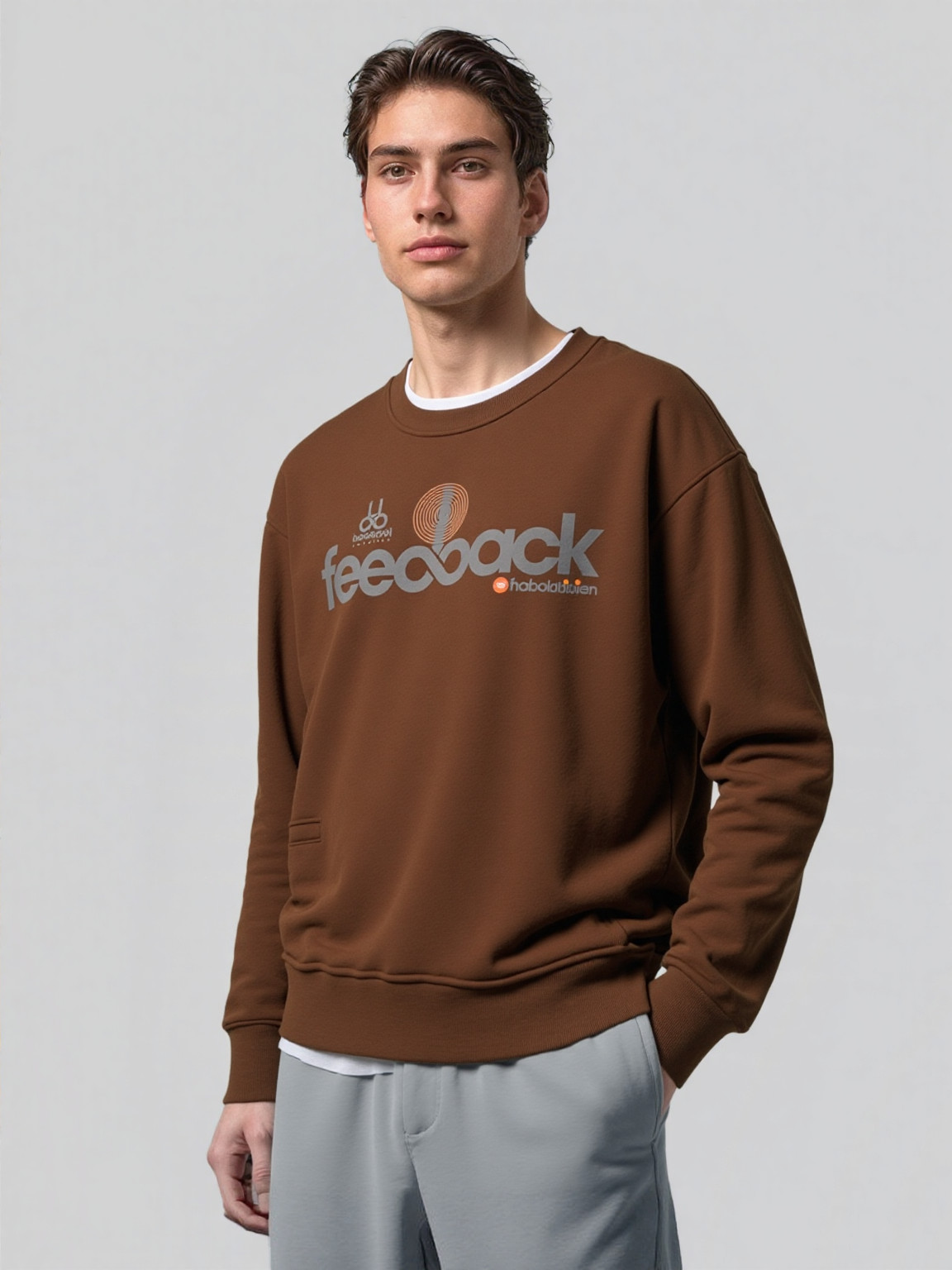 Young man wearing a brown sweatshirt with graphic text and gray pants, standing against a plain light gray background.