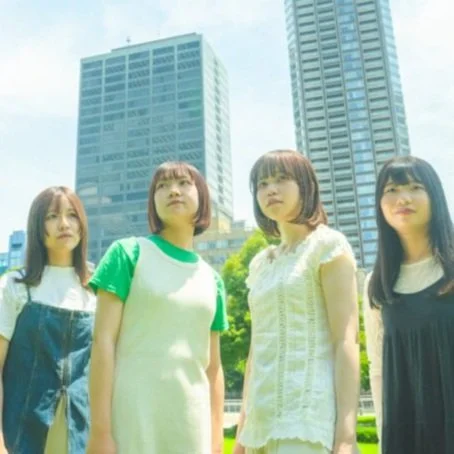 Four young women standing outdoors in an urban park with tall skyscrapers in the background, wearing casual clothing.