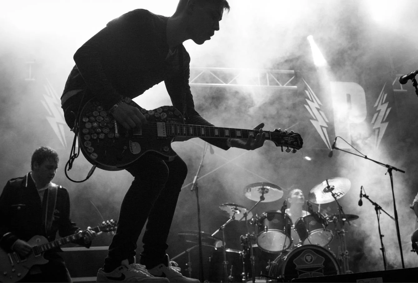 Silhouette of a band performing on stage, with a guitarist in the foreground and a drummer in the background, illuminated by stage lights and surrounded by stage equipment.