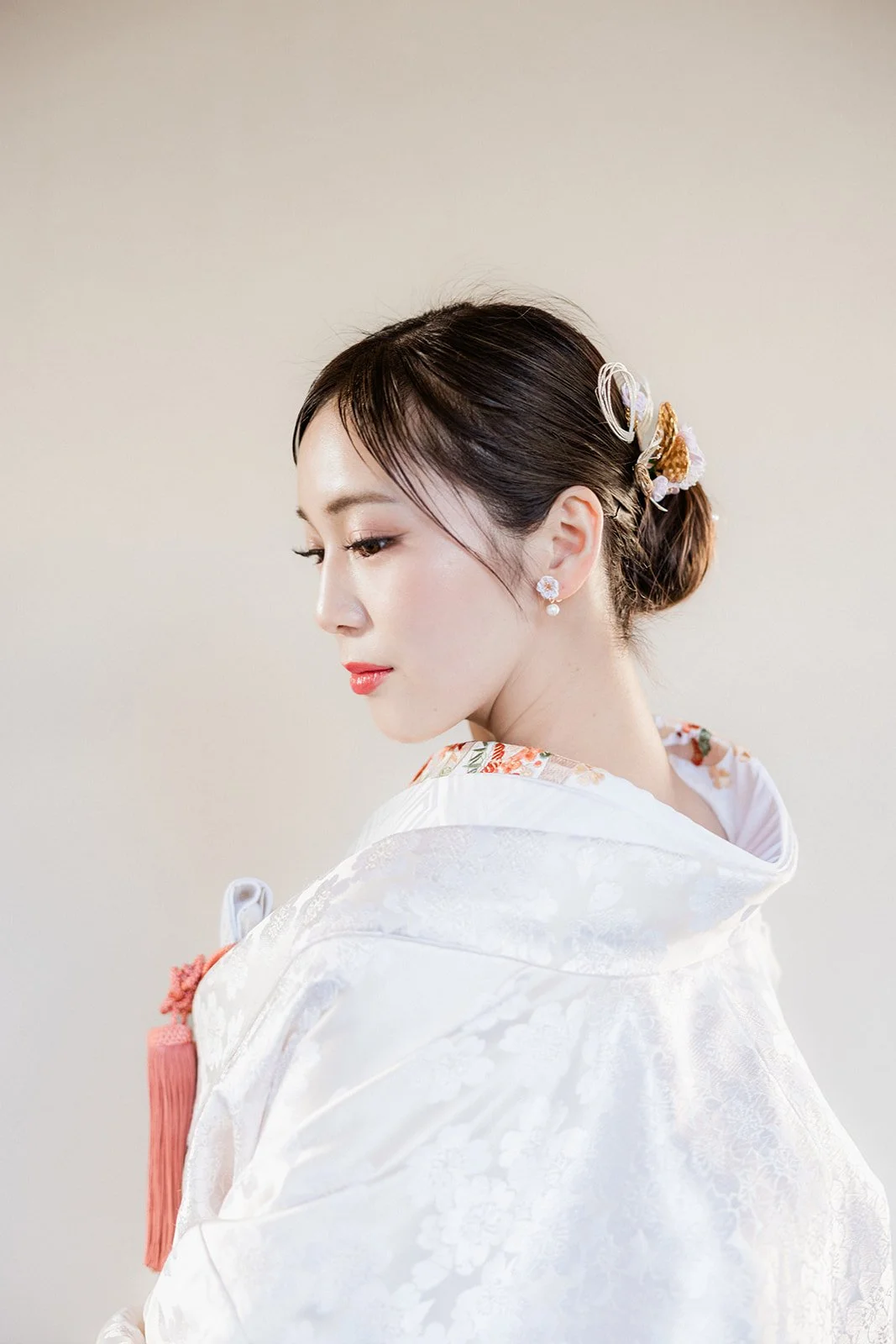 A couple dressed in traditional Japanese wedding attire standing outdoors under blooming cherry blossom trees, with traditional Japanese buildings in the background.