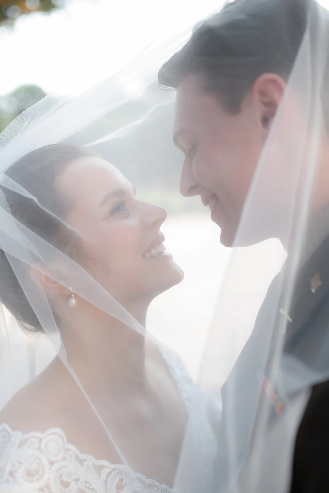 A bride and groom smiling under a sheer veil, looking into each other's eyes outdoors.