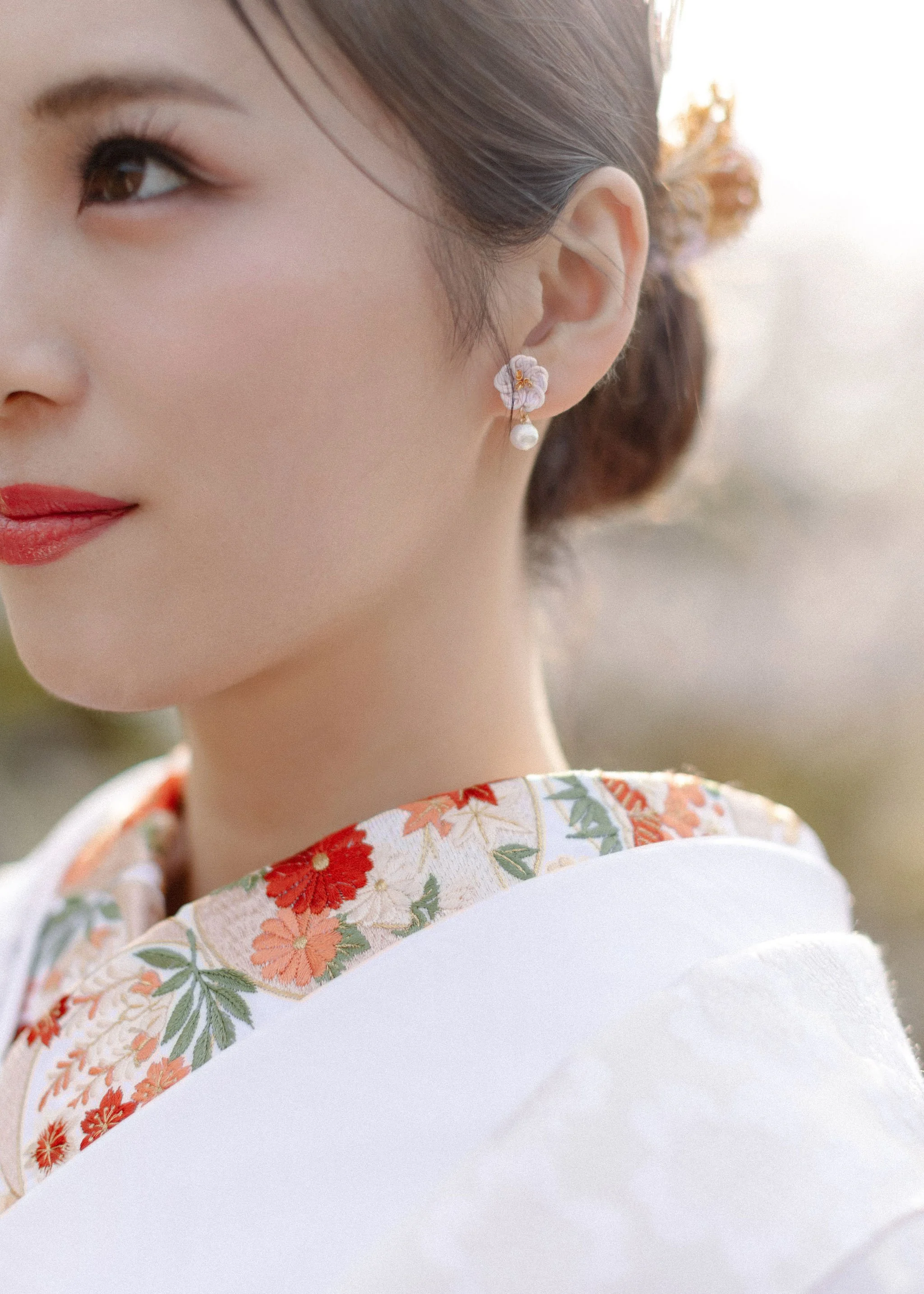 Close-up of a woman's face and shoulder, wearing a floral kimono, with earrings featuring a flower and a pearl, outdoors with soft sunlight.