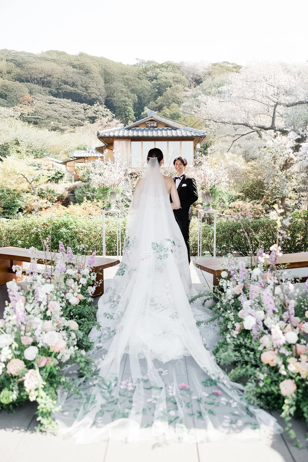 Brunette woman in a white wedding dress holds a large bouquet of pink, peach, and white flowers with greenery and a pink ribbon.