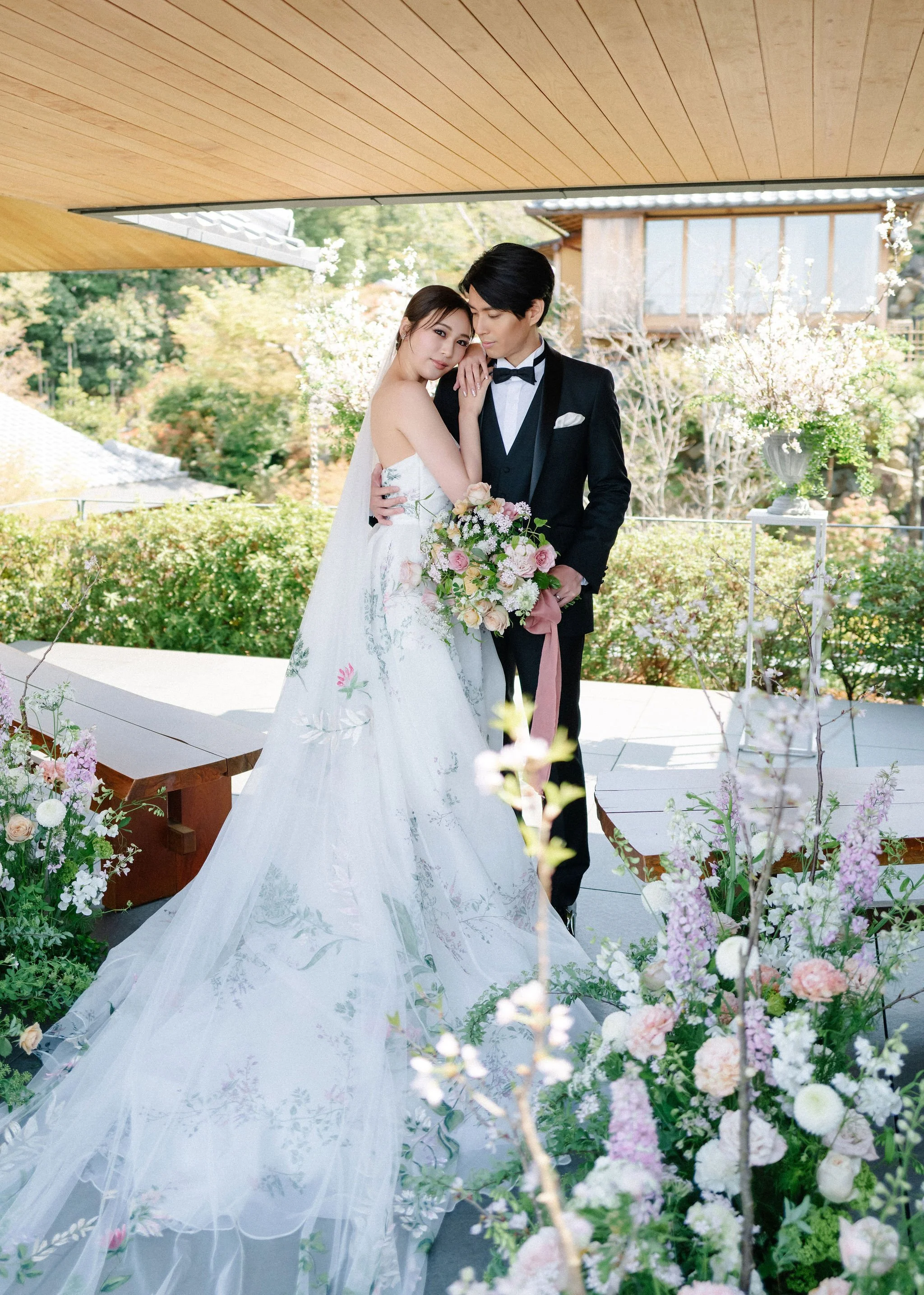 Bride and groom posing together outdoors during a wedding, with the bride holding a bouquet and wearing a white wedding gown, and the groom in a black tuxedo, surrounded by floral arrangements and greenery.