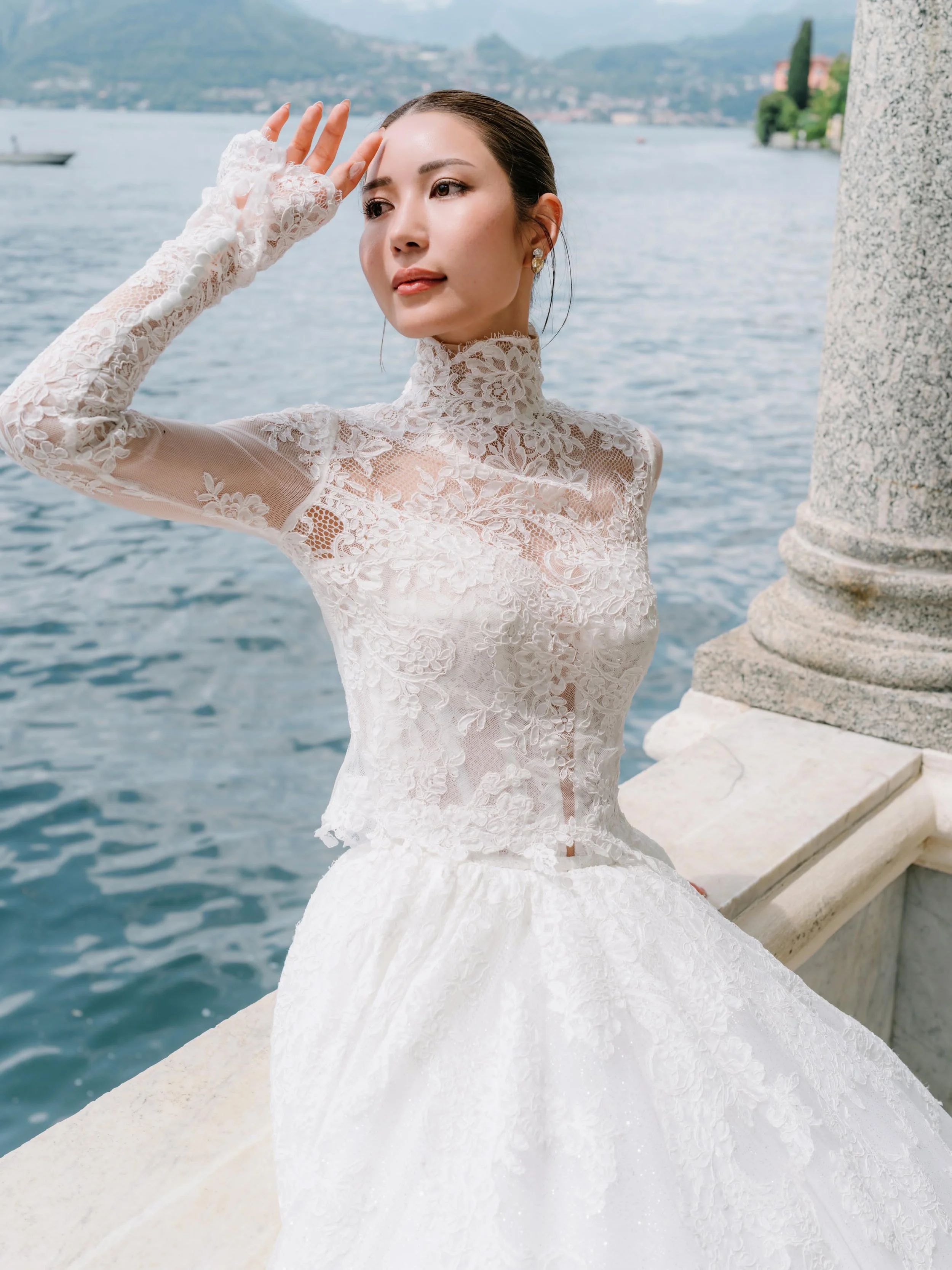 Bride in a white lace wedding dress with a veil, looking through the veil with her hands raised, outdoors, with greenery in the background.