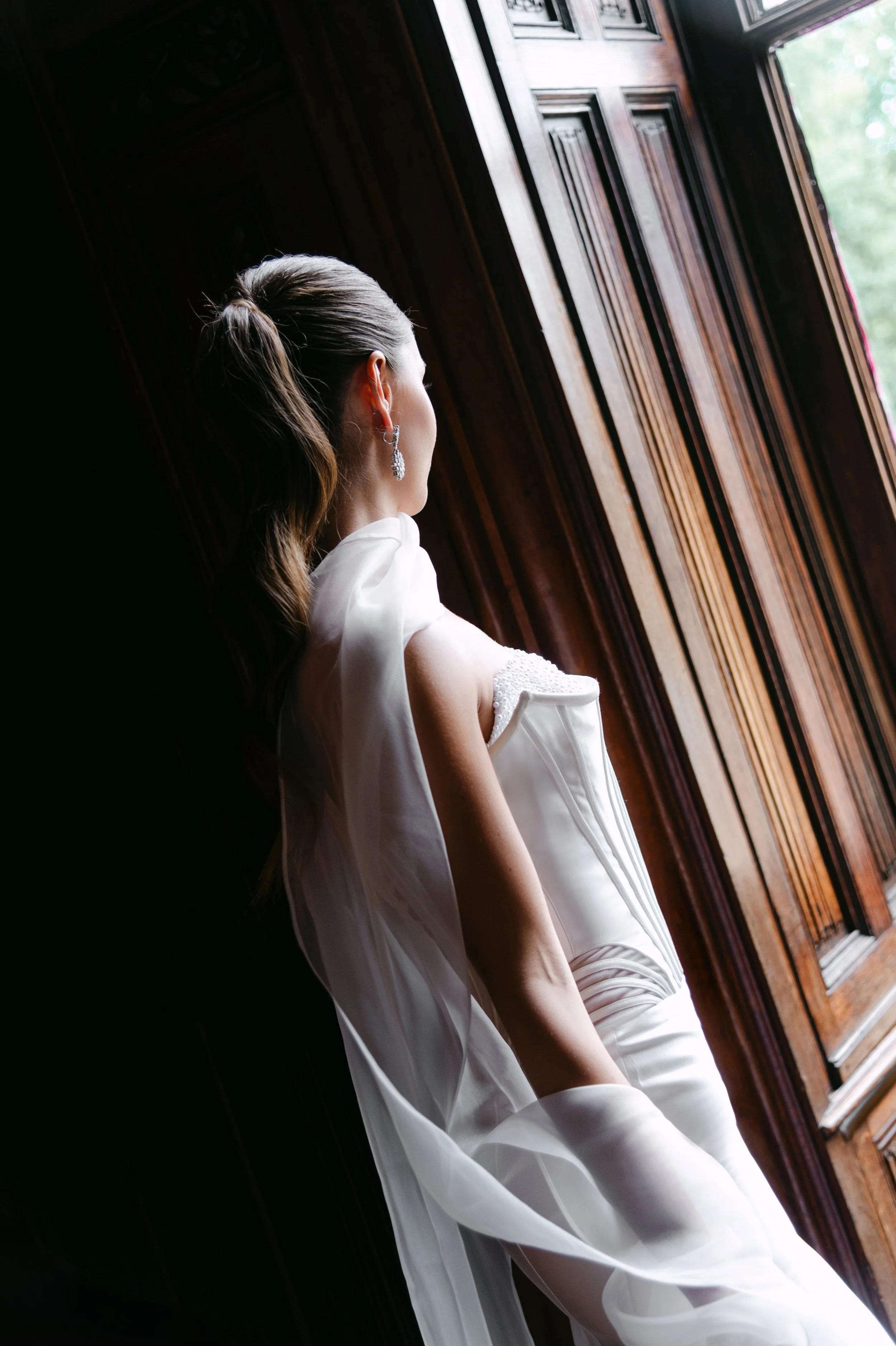 A bride in a white wedding gown standing on a city balcony, looking away from the camera, with historic buildings in the background.