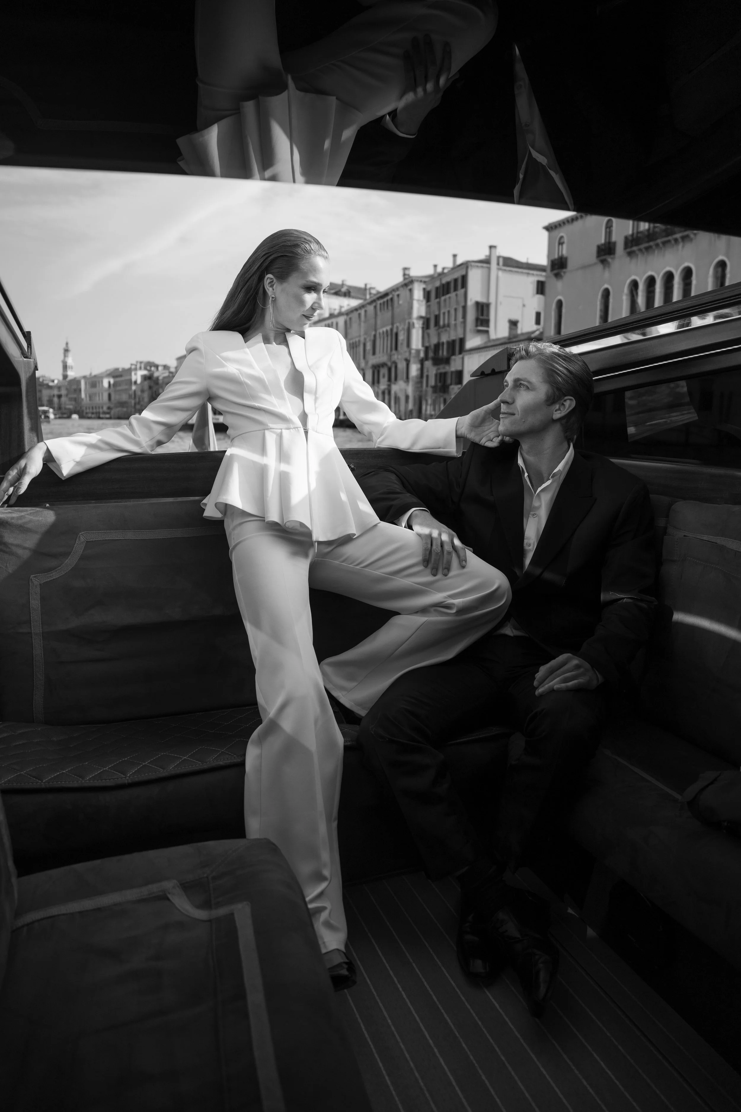A black and white photo of a woman and a man inside a boat. The woman is standing with her leg on the seat, wearing a stylish white suit, and the man is sitting, looking at her, in a dark suit. They are in a city with buildings visible through the boat window.