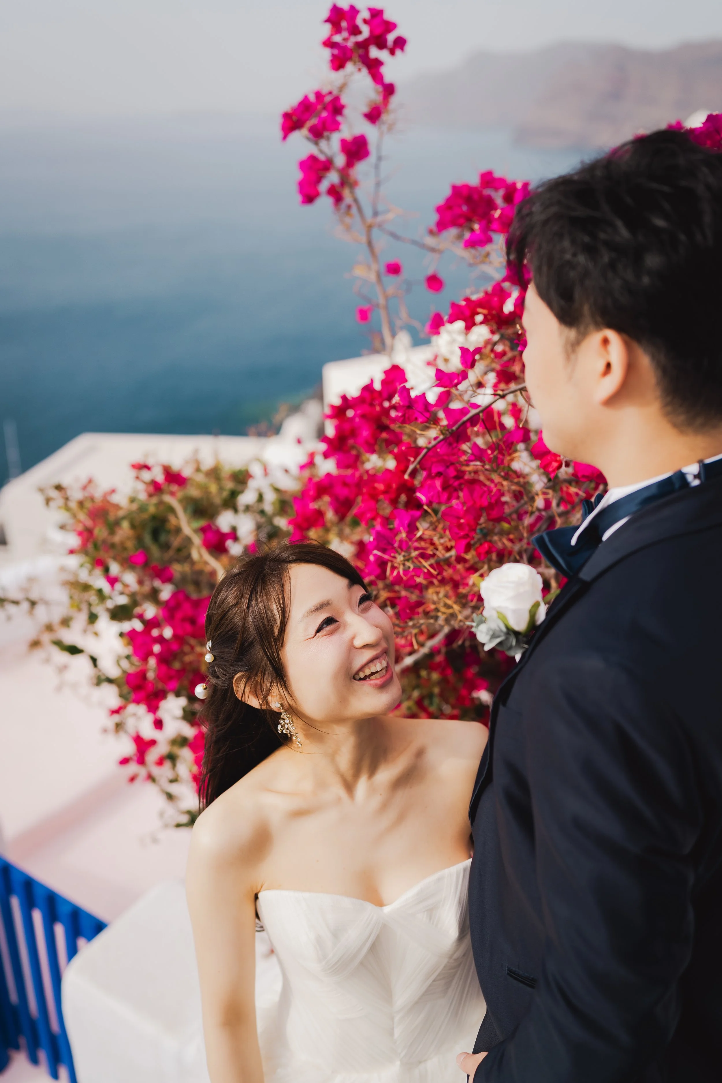 A bride and groom embrace during their wedding, with bright pink bougainvillea flowers and the sea in the background.