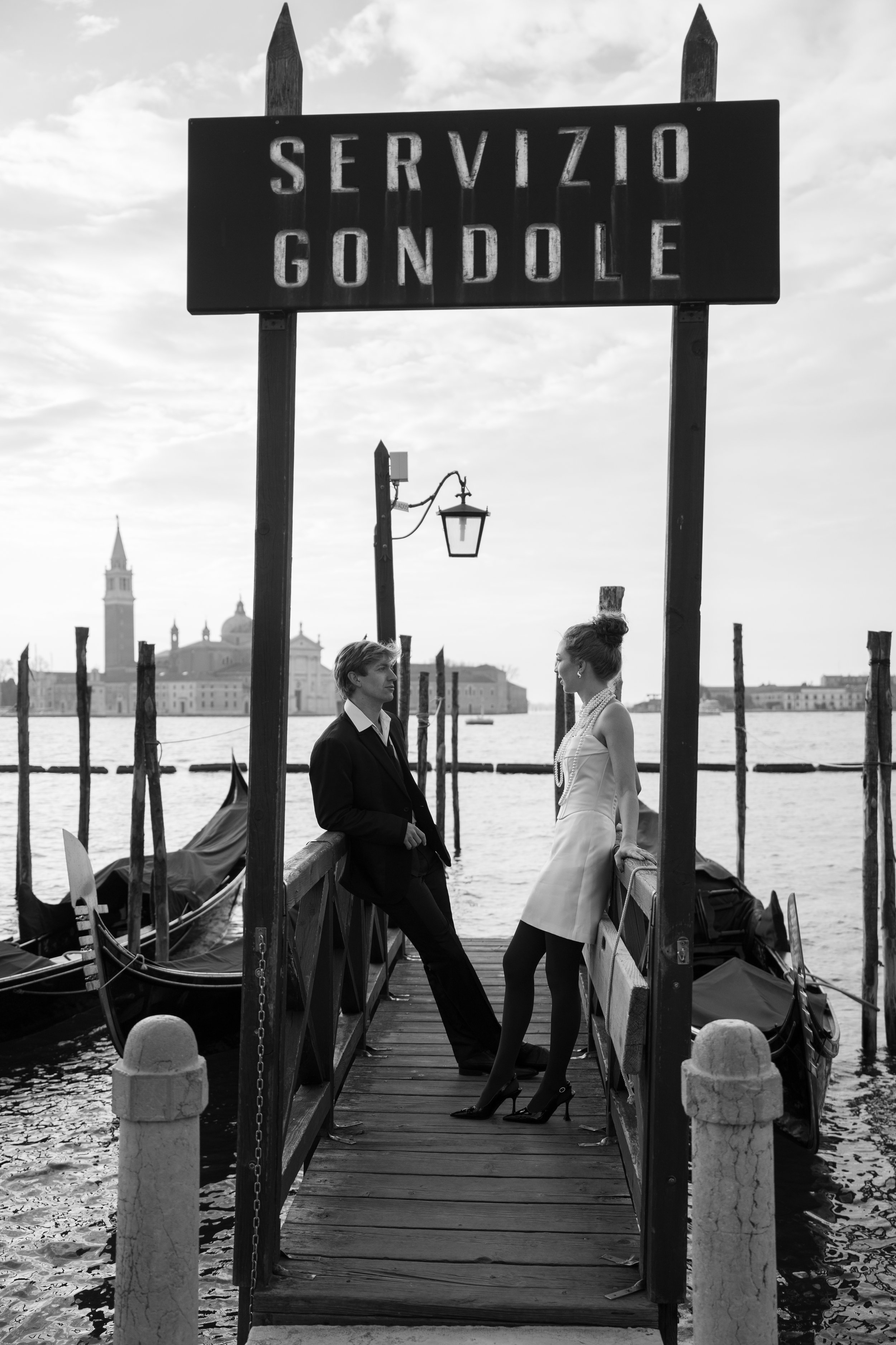 A black and white photo of a gondola dock in Venice, Italy, with a man and woman dressed in vintage clothing talking on the dock.