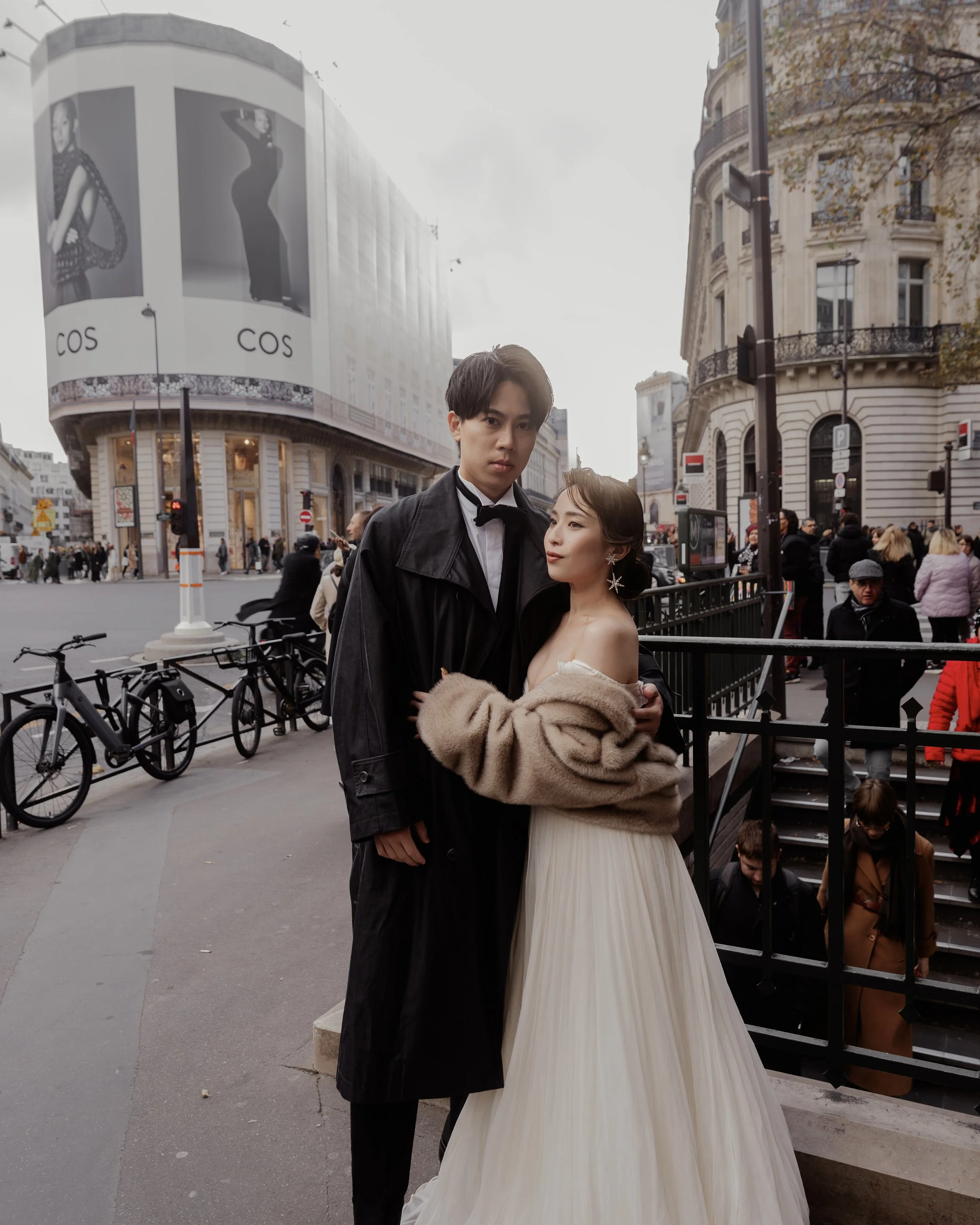 A romantic couple stands close together outside the Louvre Museum in Paris, with the glass pyramid visible in the background.
