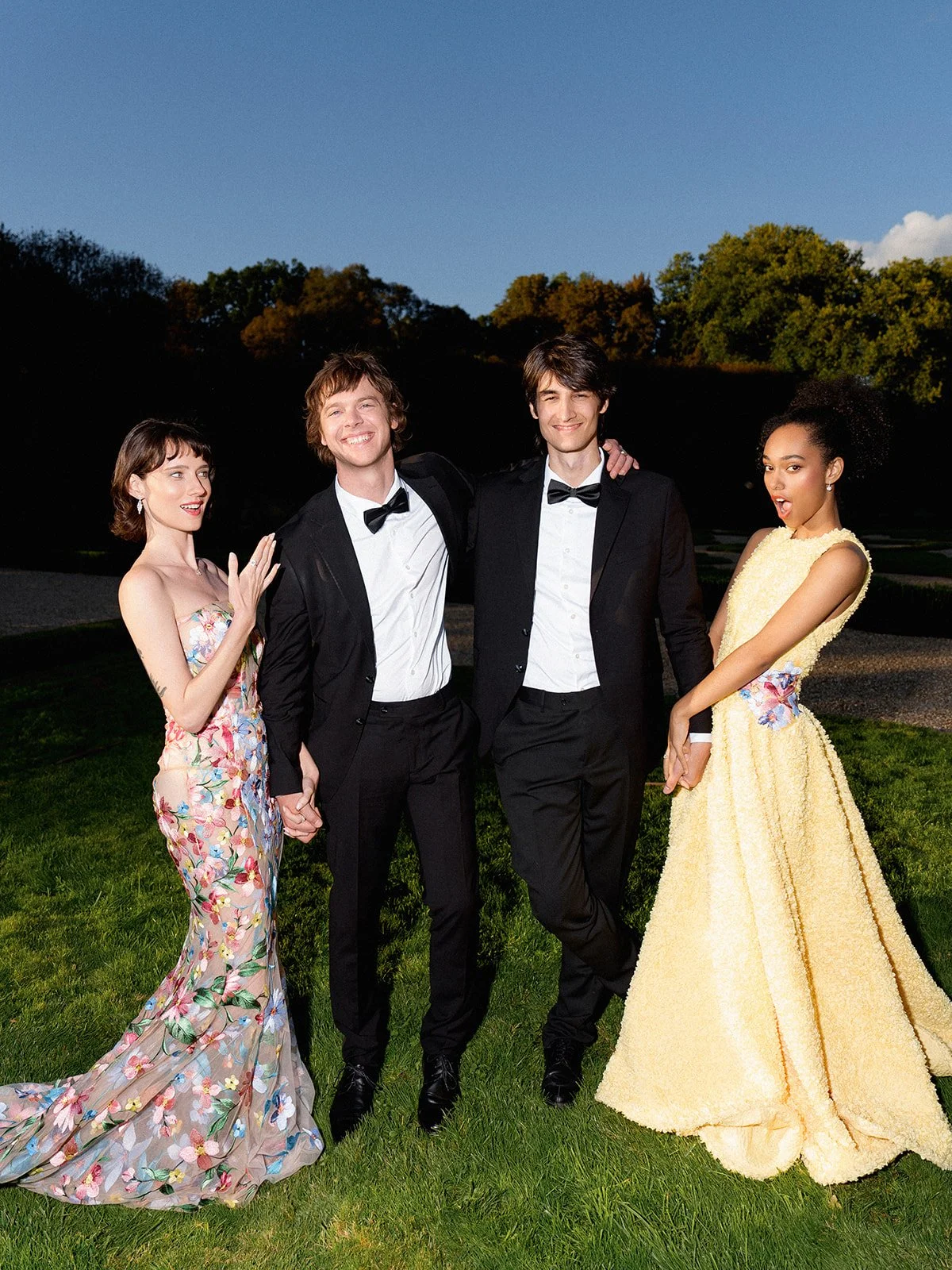 Four young adults dressed in formal attire standing outdoors on grass during evening, with trees and a clear sky in the background.