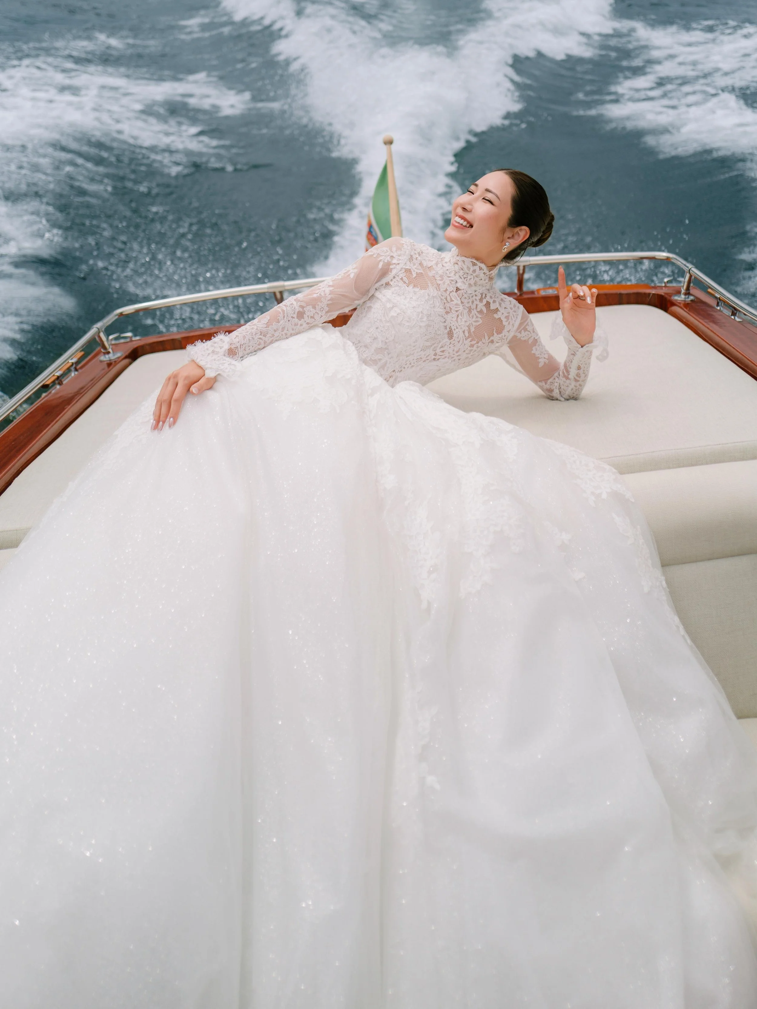 A woman in a white lace dress by the water with mountains in the background.