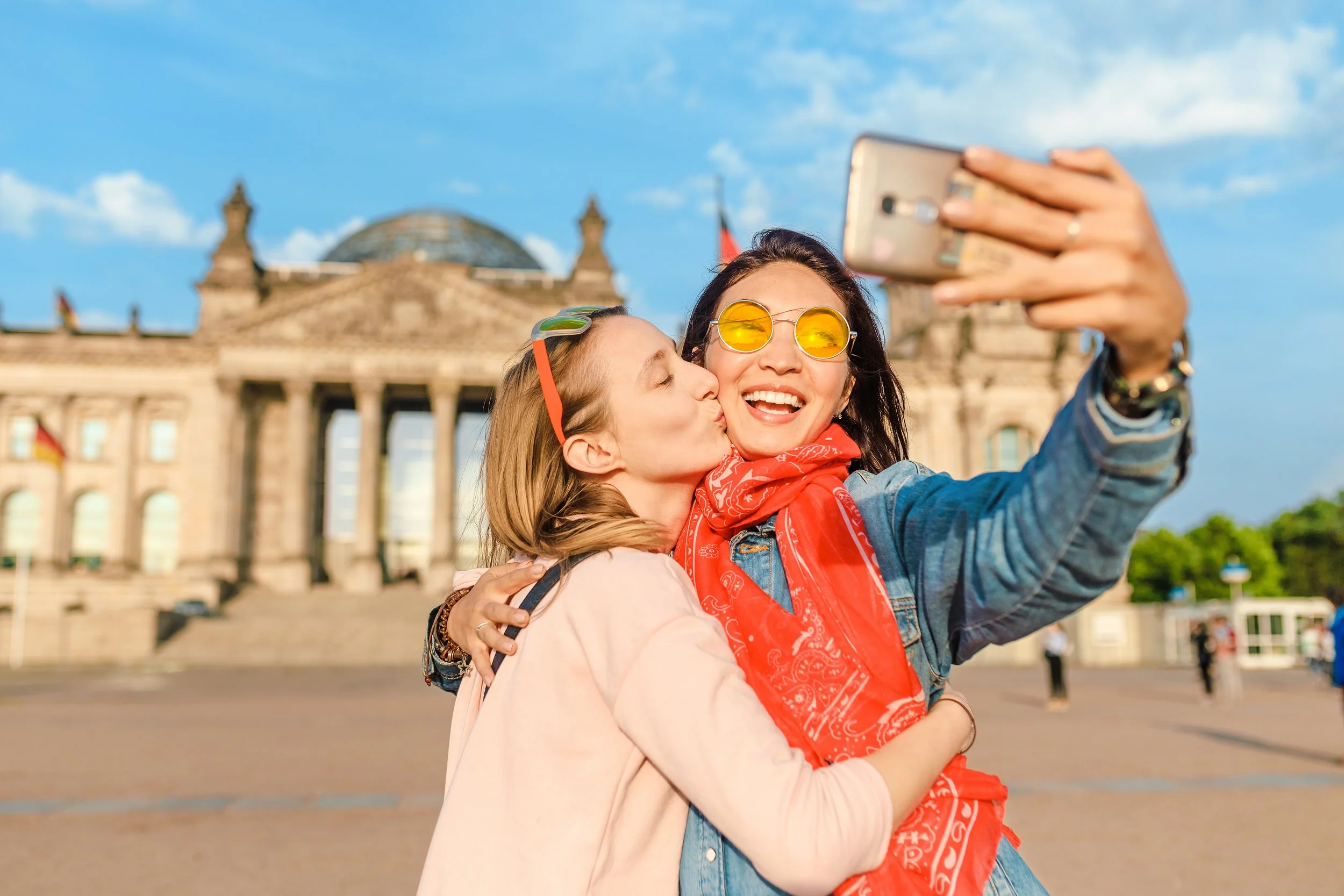 Two women take a selfie in front of the Reichstag building in Berlin. One woman kisses the other on the cheek.