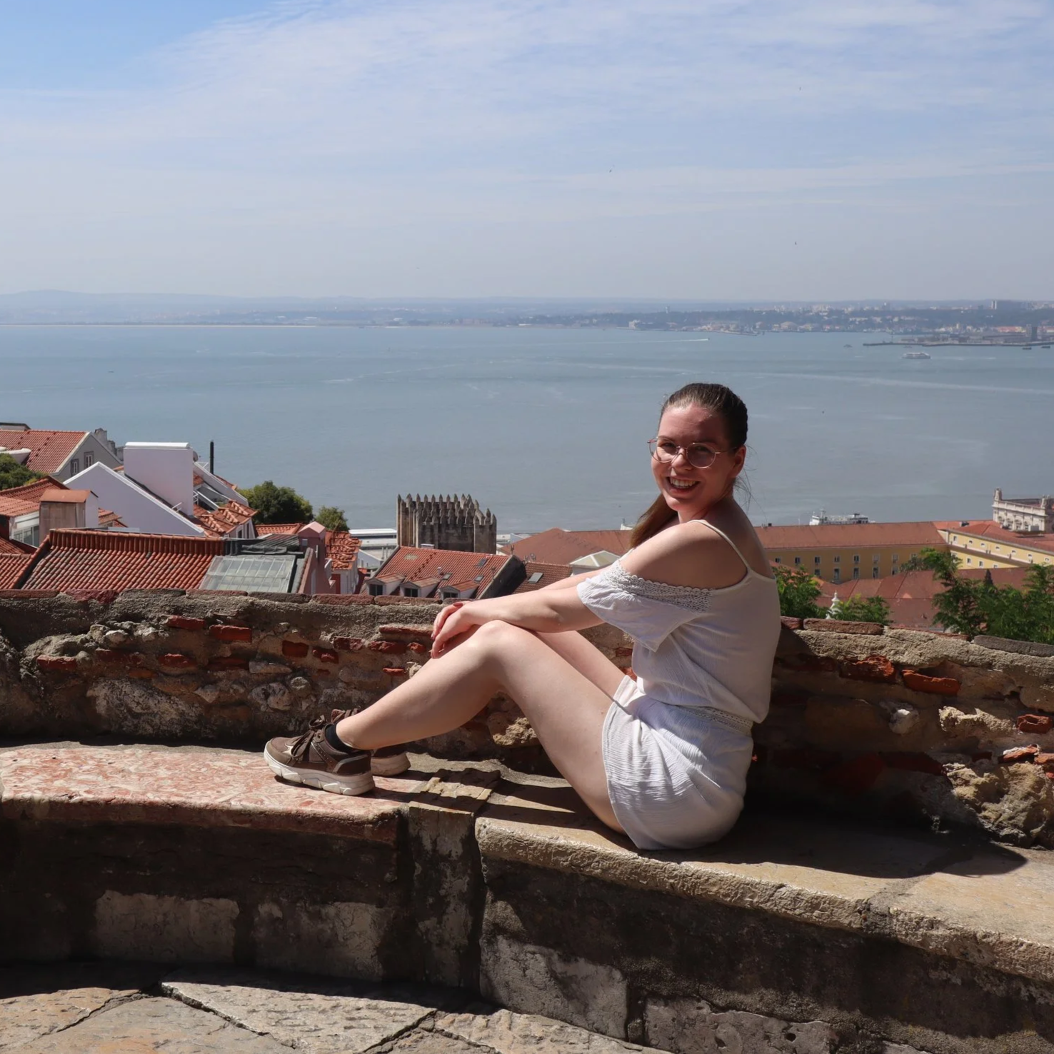 Young woman sitting on a stone wall, looking laughing into the camera with water and city in the background.