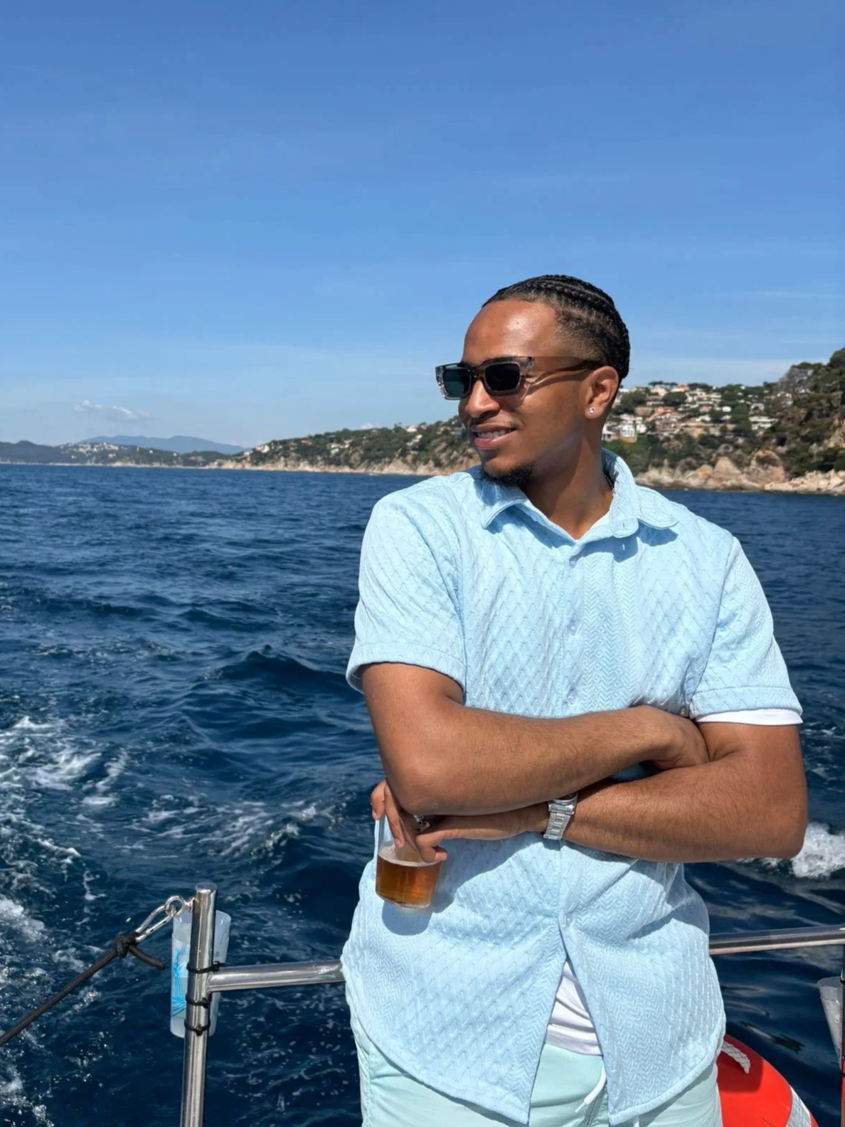 A man with sunglasses and short hair stands on a boat and looks into the distance, in the background the sea and a coastline with houses under a sunny sky.