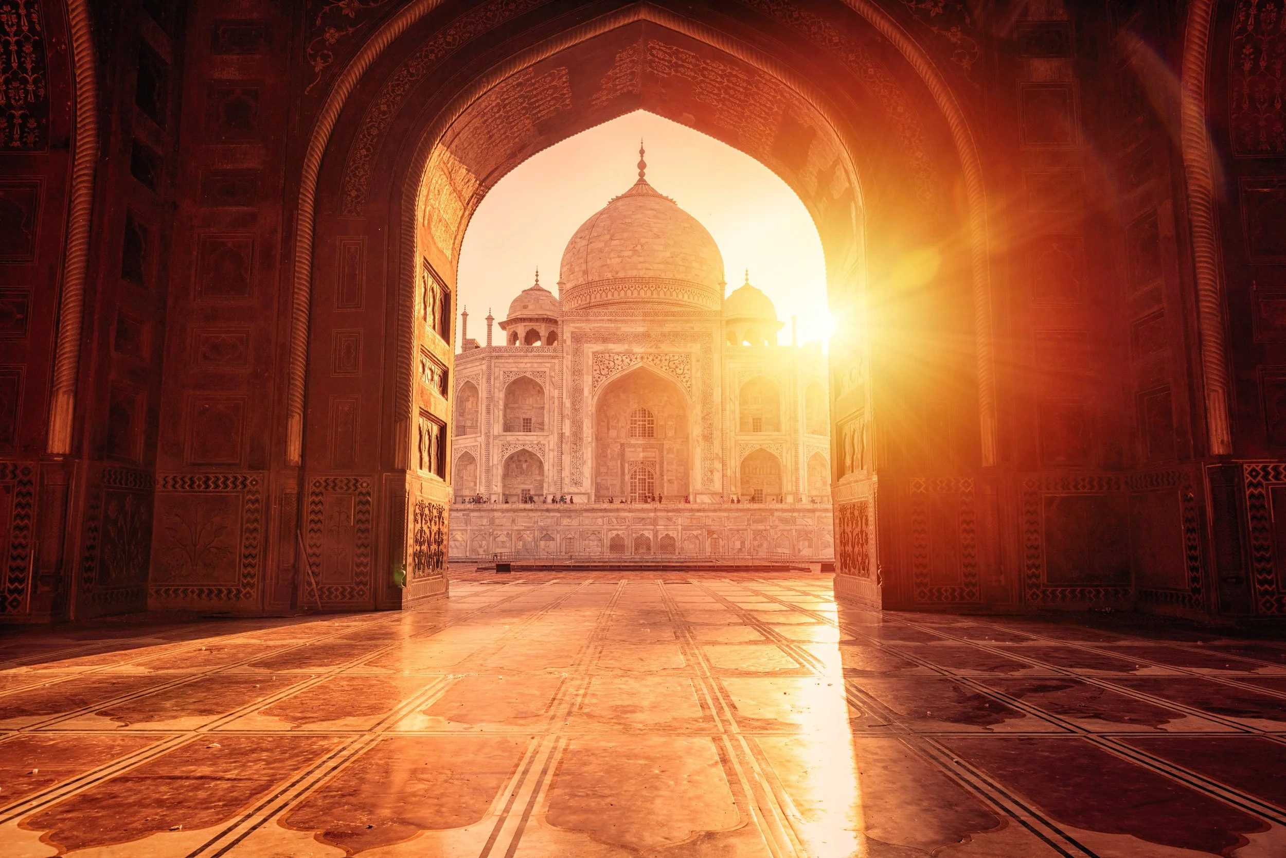 A view through an archway of the Taj Mahal at sunrise, with warm lighting effects and detailed patterns on the floor and walls.