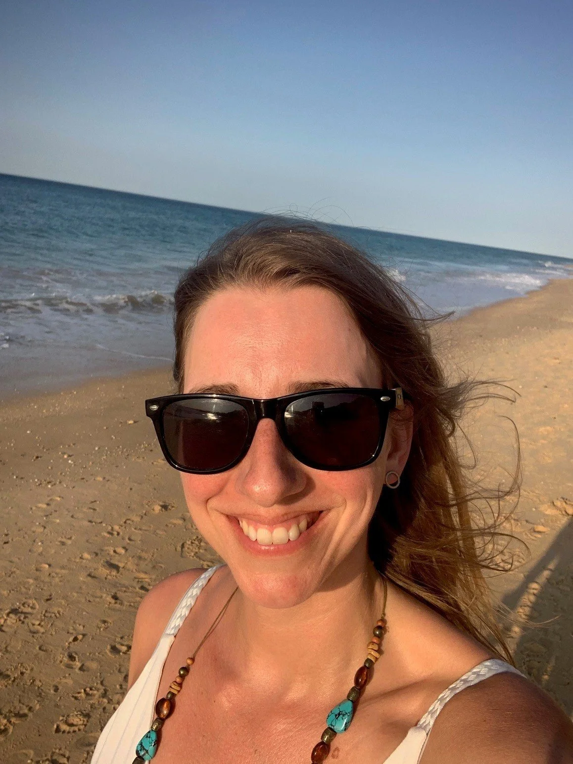 Woman with sunglasses on the beach, sunset in the background, smiling.