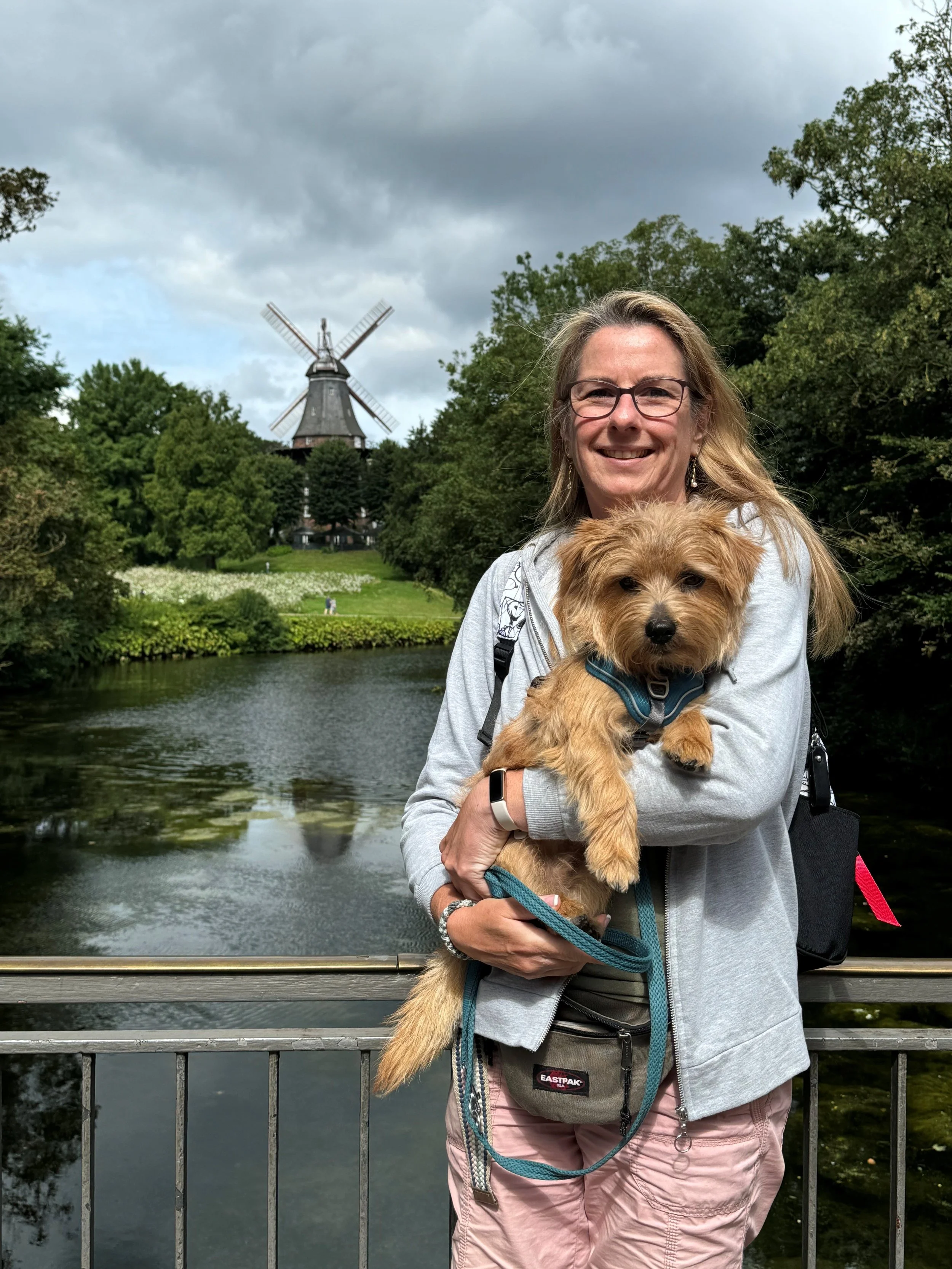 Maja holds her dog on a bridge, with water and trees in the background, and a windmill under a cloudy sky.