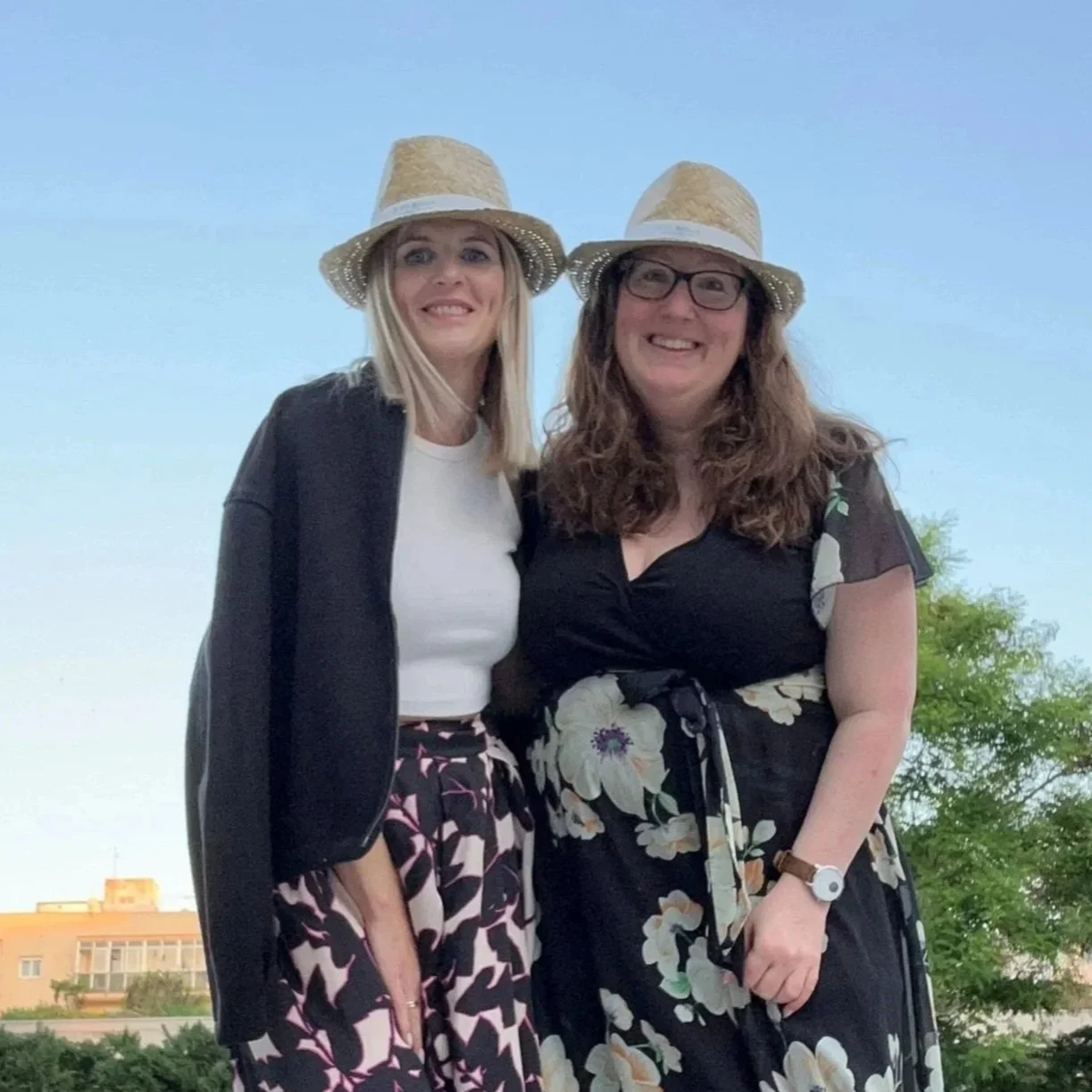 Two women in summer dresses with straw hats, smiling and posing outside in the evening.