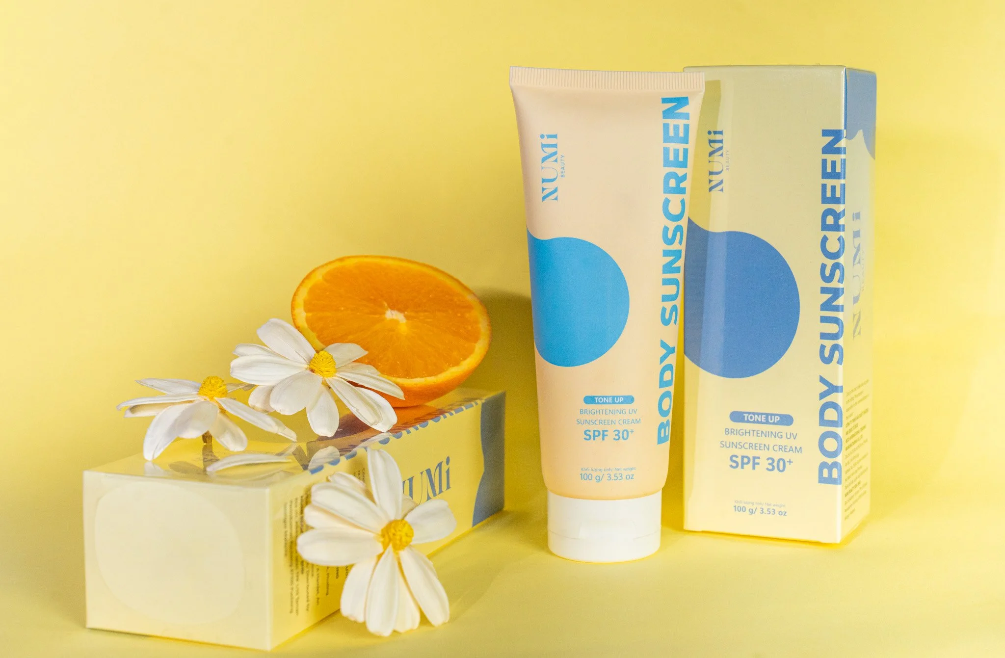 A yellow background featuring skincare products, an orange half, and white chamomile flowers with yellow centers. The skincare item is a tube and box labeled 'BODY SURF CREAM SPF 30+' in blue and yellow colors.
