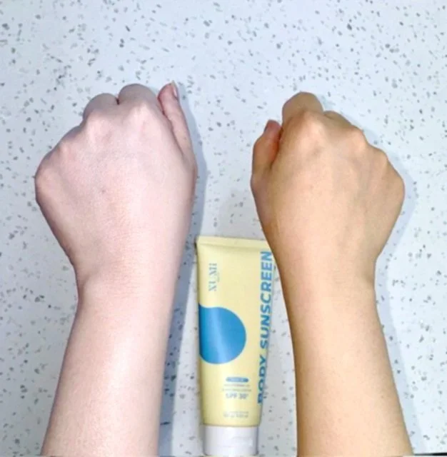 Two clenched fists, one with light skin and one with darker skin, placed side by side on a speckled surface with a tube of SPF 50 sunscreen between them.
