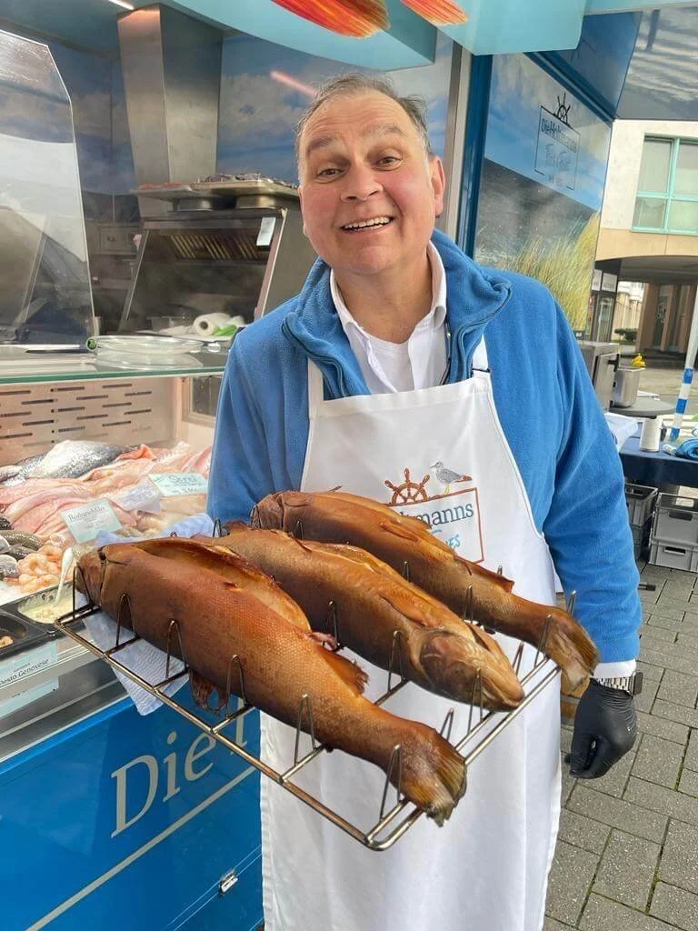 Ein Mann trägt eine Schürze und präsentiert drei geräucherte Fische auf einem Rost.