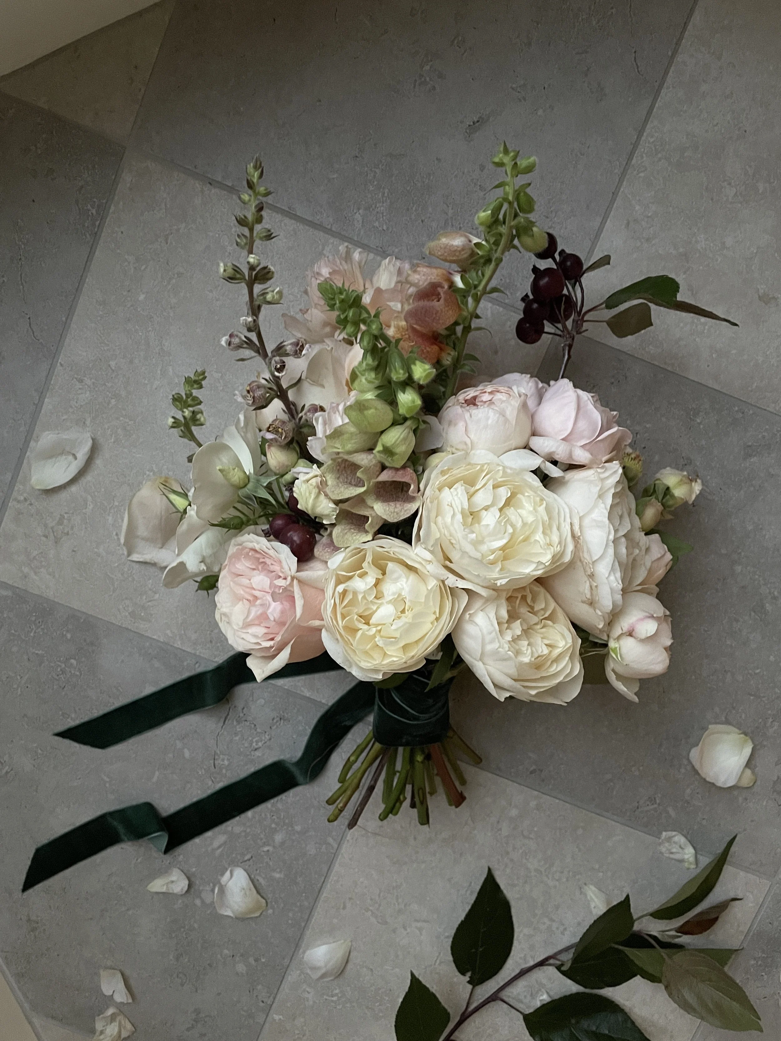 A bouquet of pale pink and white roses, with accents of greenery and dark red berries, resting on a tiled floor with some scattered white flower petals nearby.