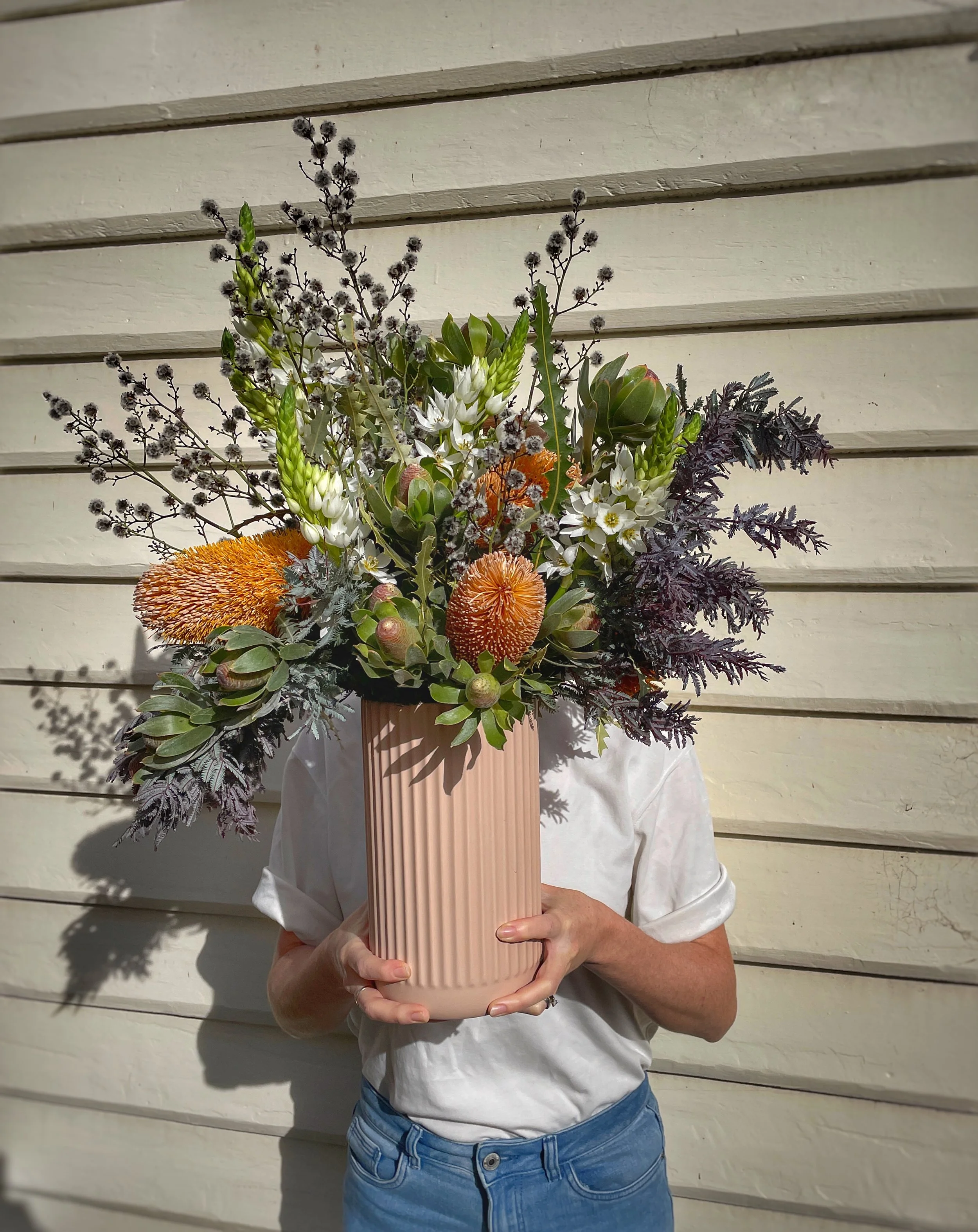 Person holding a large pink vase filled with an assortment of white, orange, and purple flowers and greenery, in front of a weatherboard house.
