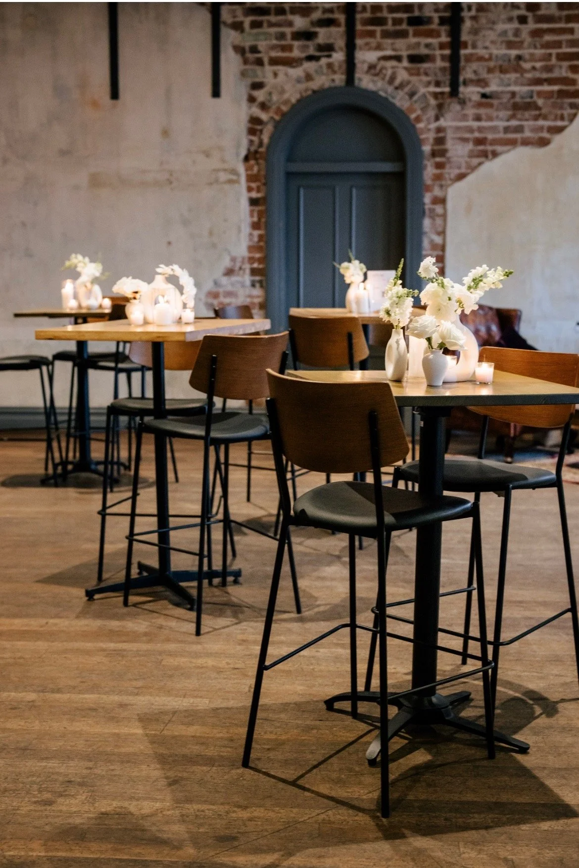 A wedding reception with wooden tables, black chairs, and white flower arrangements in vases, with candles. Exposed brick wall and a blue arched door in the background.