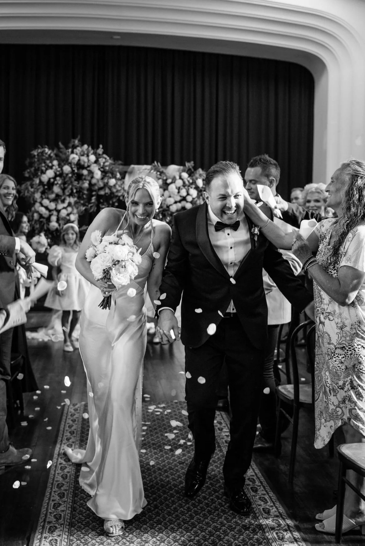Black and white photo of a wedding celebration with a bride in a long dress holding a bouquet and a groom in a tuxedo laughing as they walk hand-in-hand through a confetti shower, surrounded by friends and family.