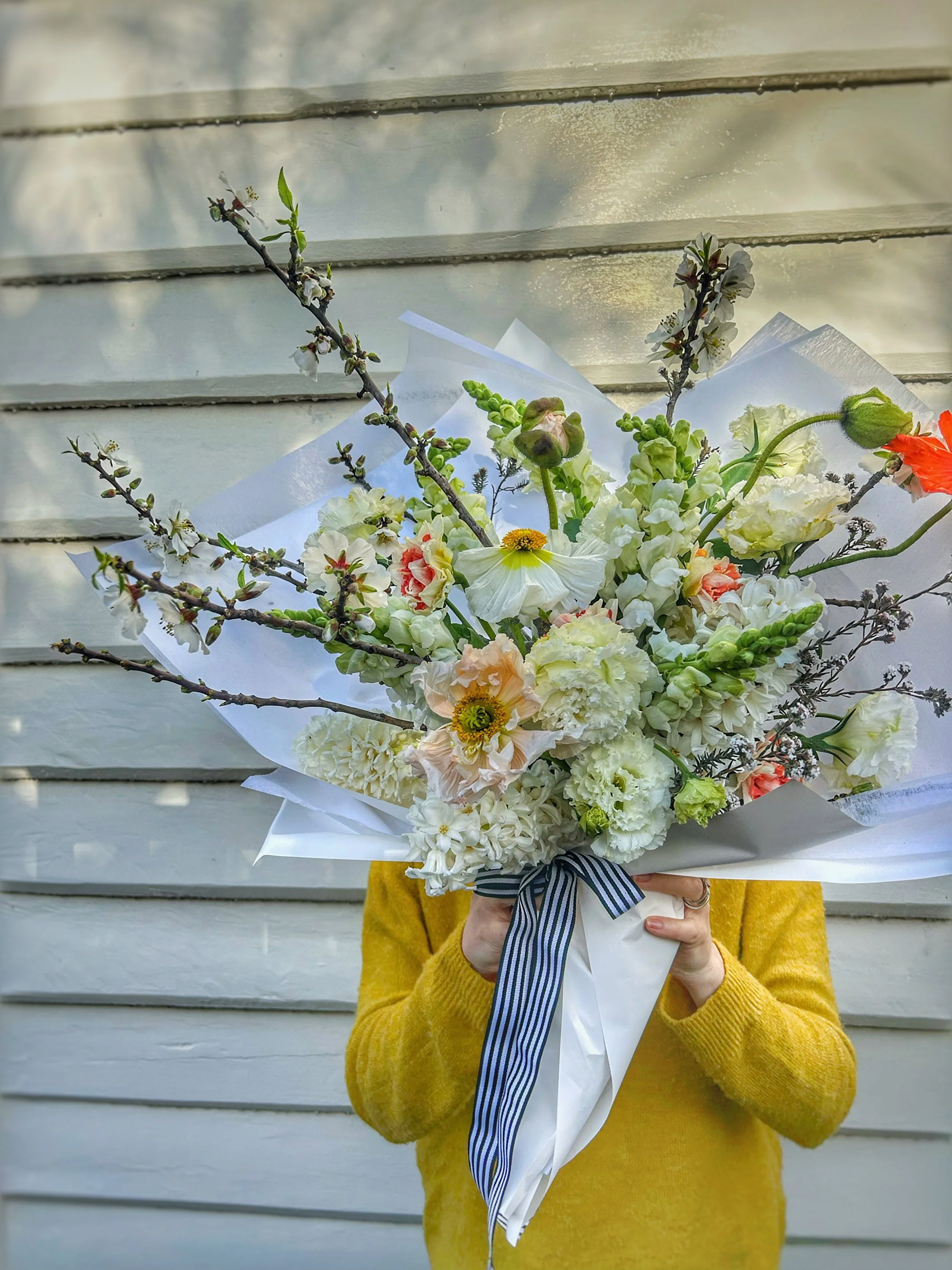 Person in a yellow sweater holding a large bouquet of white and light pink flowers in front of a weatherboard house.