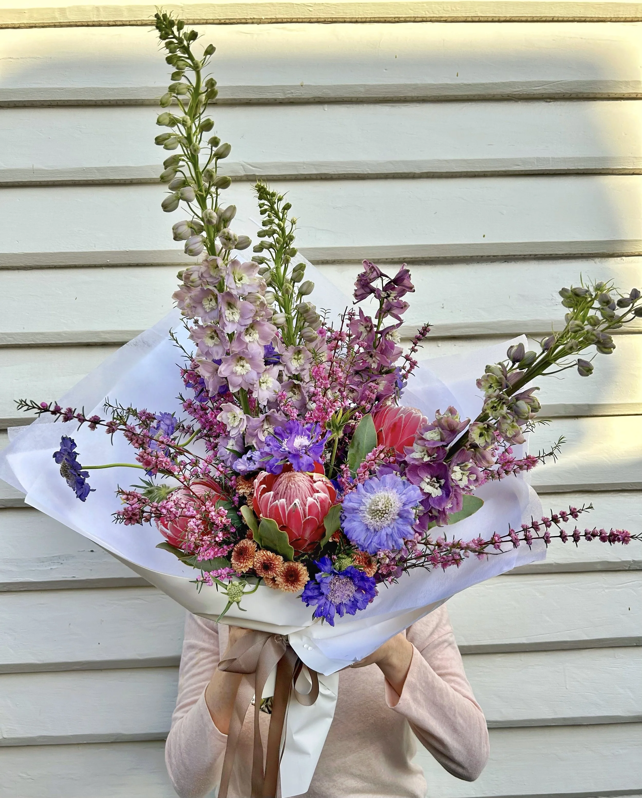 Person holding a large colorful bouquet of various flowers against a weatherboard house.