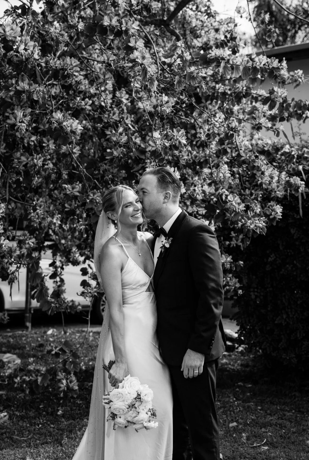 A black-and-white photo of a bride and groom standing close together outdoors in front of a large tree, smiling and sharing an intimate moment on their wedding day.