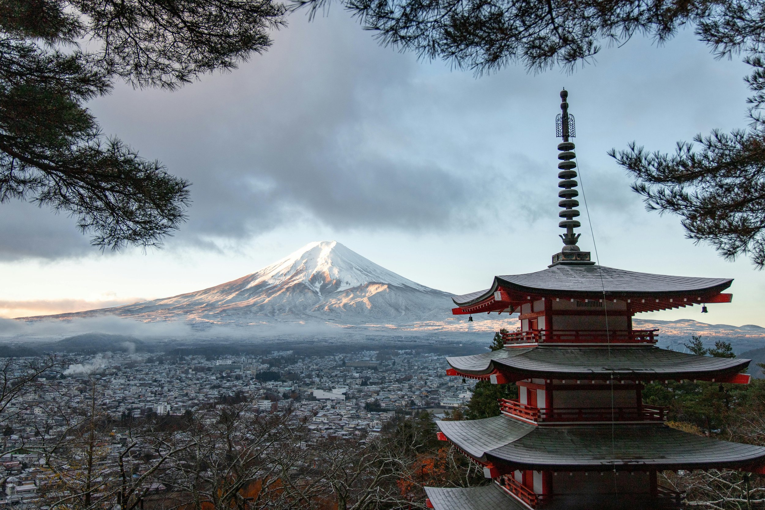 A traditional Japanese pagoda with Mount Fuji in the background, snow-capped and partially cloudy sky, taken during early morning or late afternoon.