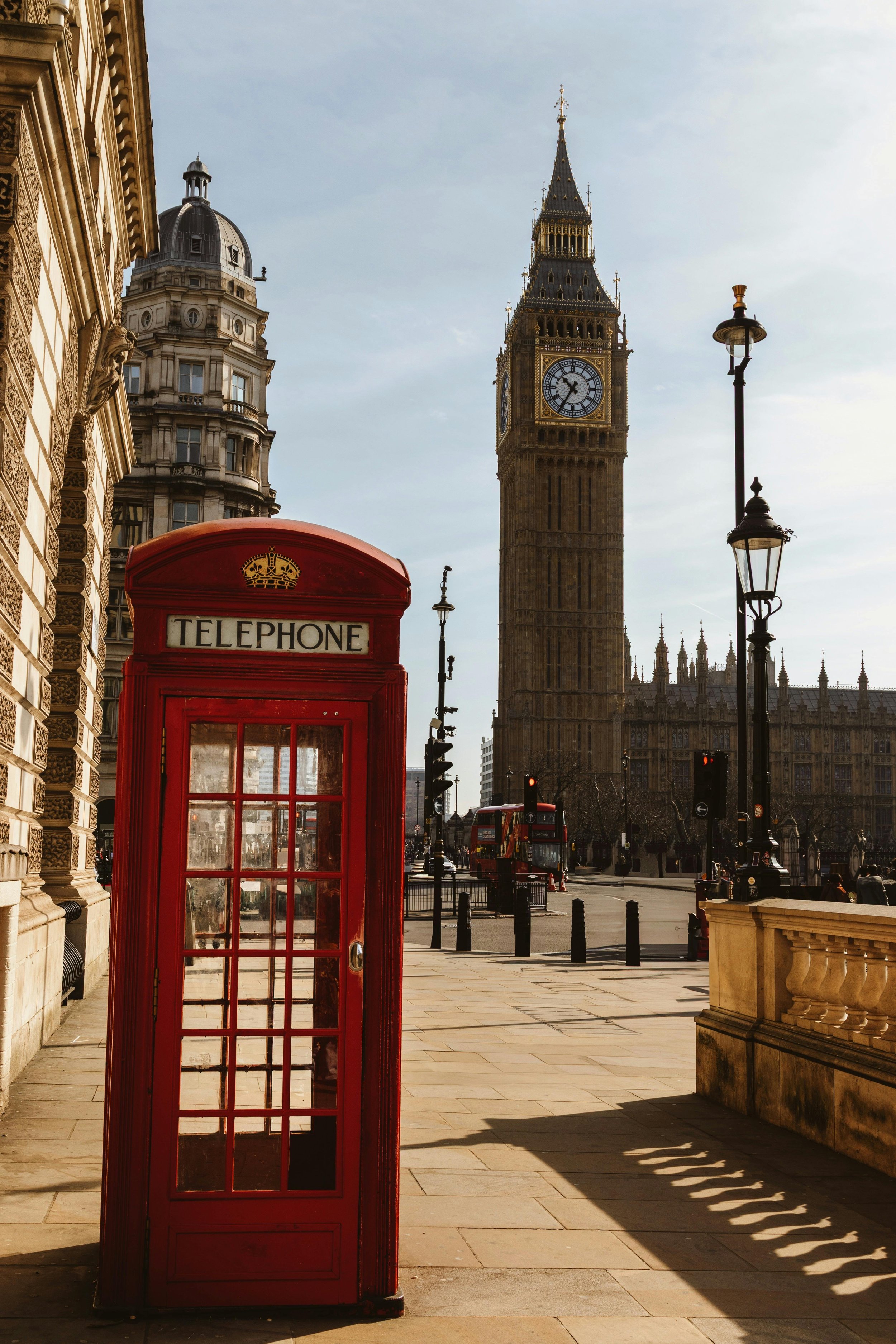 Red telephone booth on a sidewalk with Big Ben clock tower and historic buildings in London, UK in the background.
