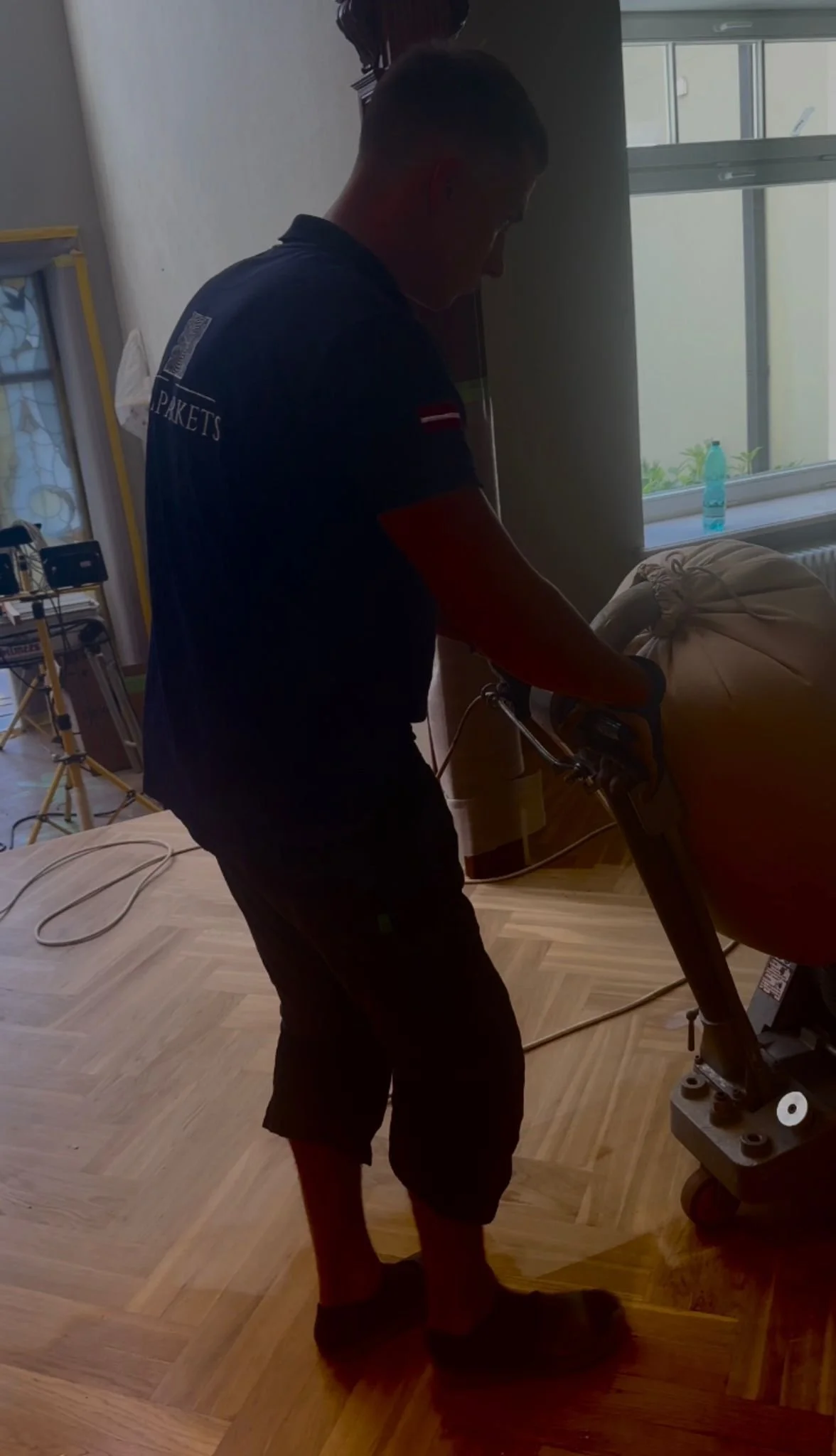 A person operating a vacuum cleaner or carpet cleaner machine in a room with wooden flooring, near a window with a water bottle on the windowsill.