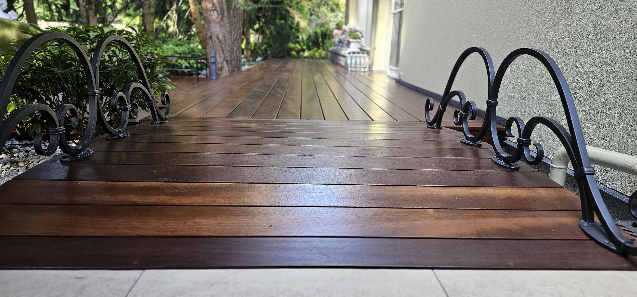 A wooden deck with polished planks and curly black metal railings, surrounded by lush greenery and plants beside a house exterior wall.