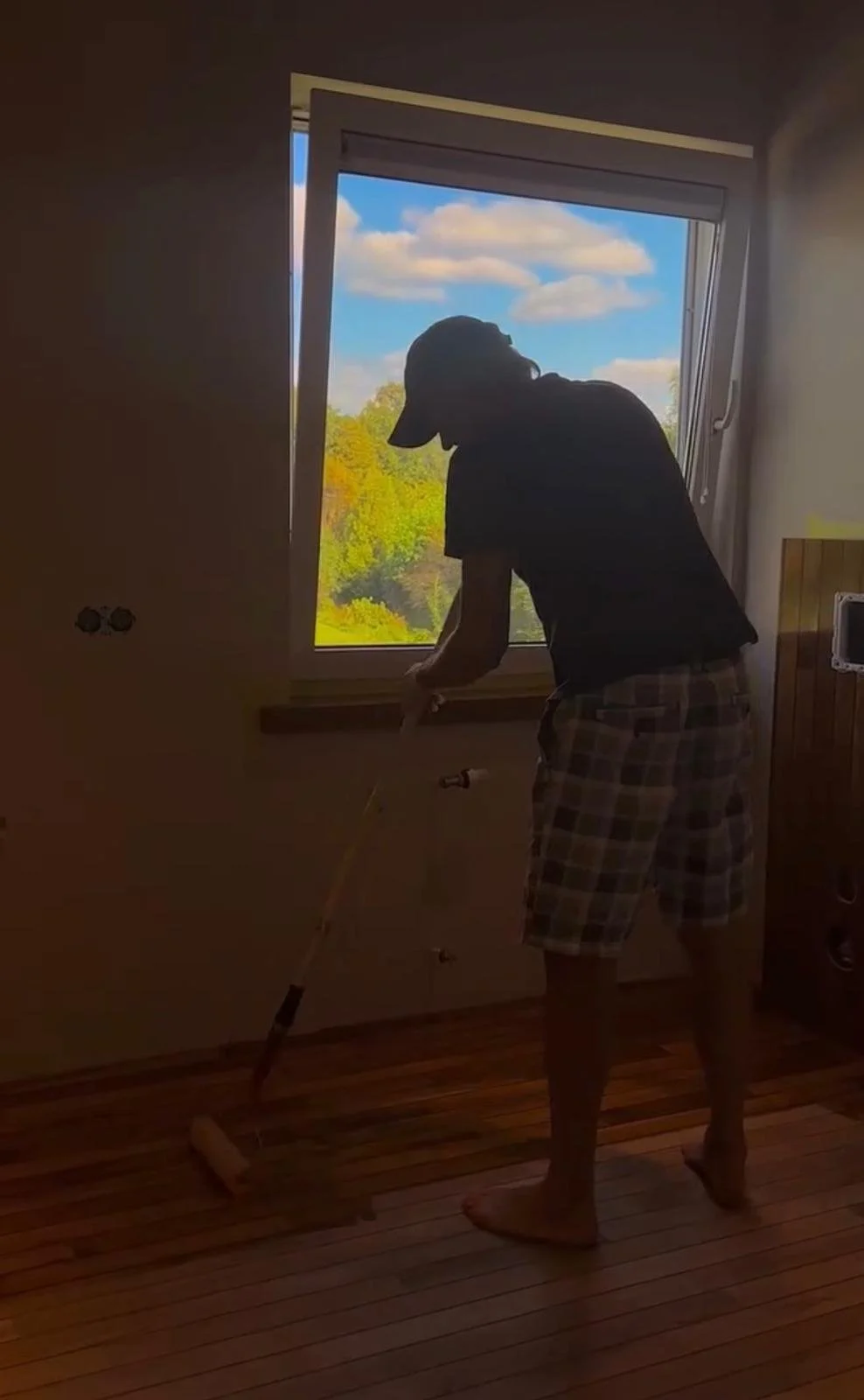 A person standing indoors by a window, wearing a cap and plaid shorts, measuring flooring with a tool, with a view of trees and a partly cloudy sky outside.