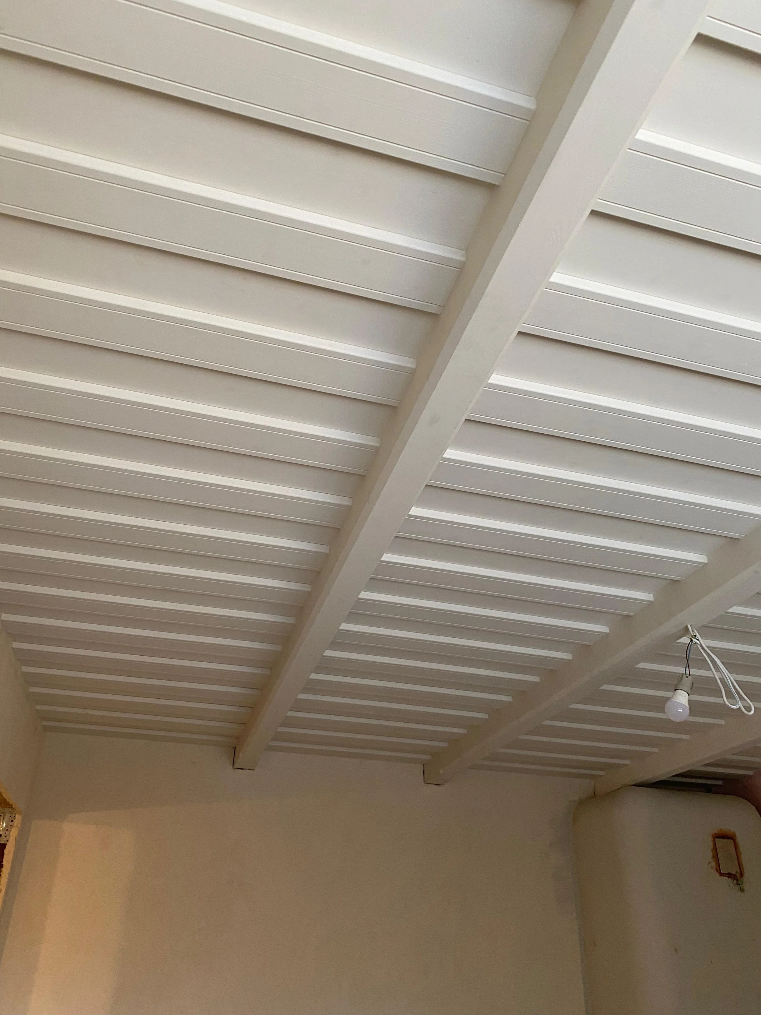Photo of a white slatted ceiling with visible beams and a hanging light bulb in a room with beige walls.