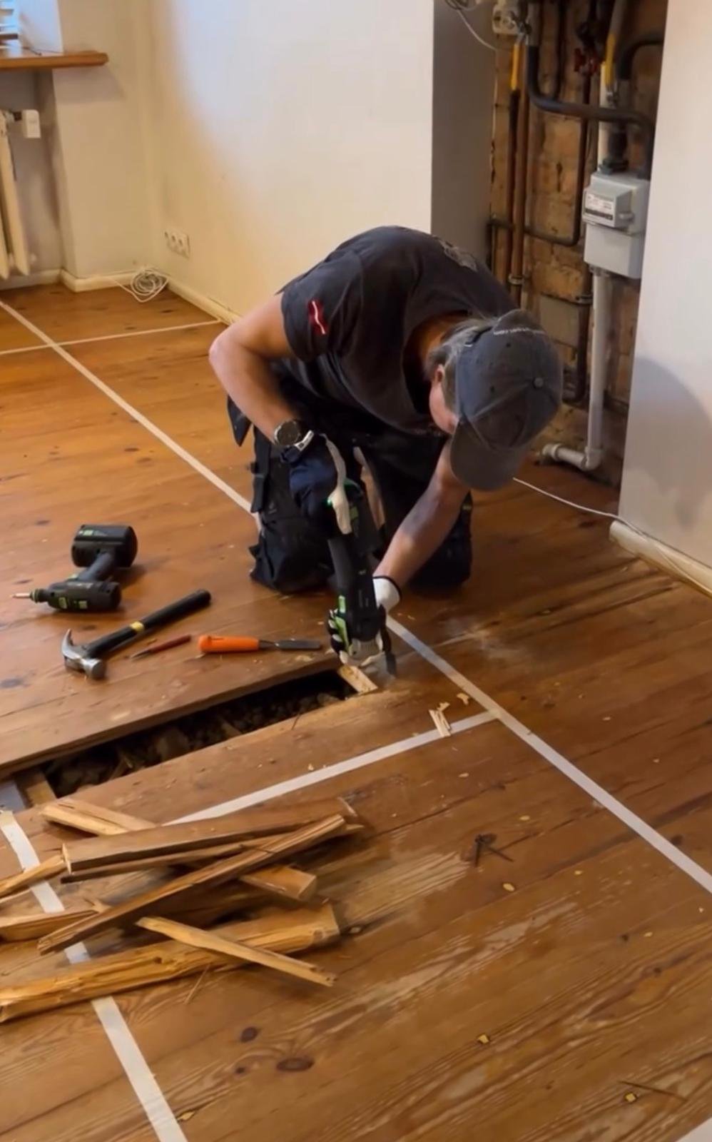 A person crouching on a wooden floor using a power drill during flooring installation or repair, with tools and wood scraps nearby.