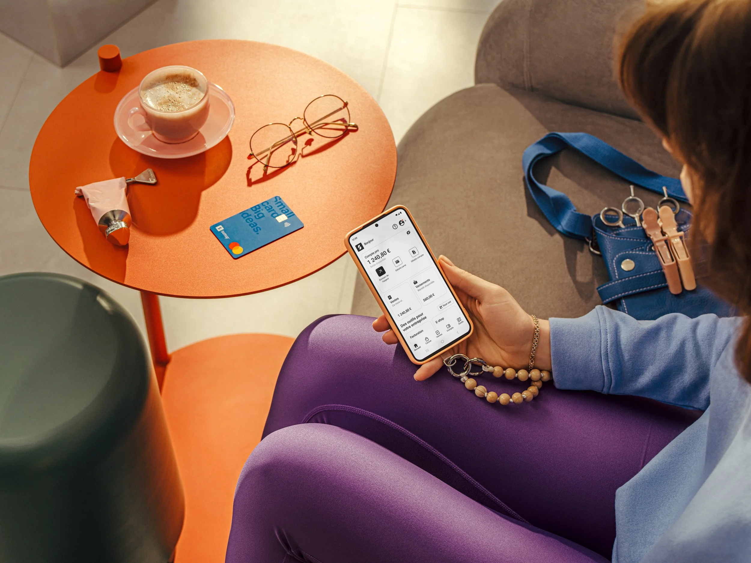 A woman sitting on a gray couch at an orange table, using a smartphone with a digital banking app. On the table, there is a cup of coffee, a tube of lipstick, sunglasses, a blue credit card, and a small orange container. A blue handbag with jewelry h