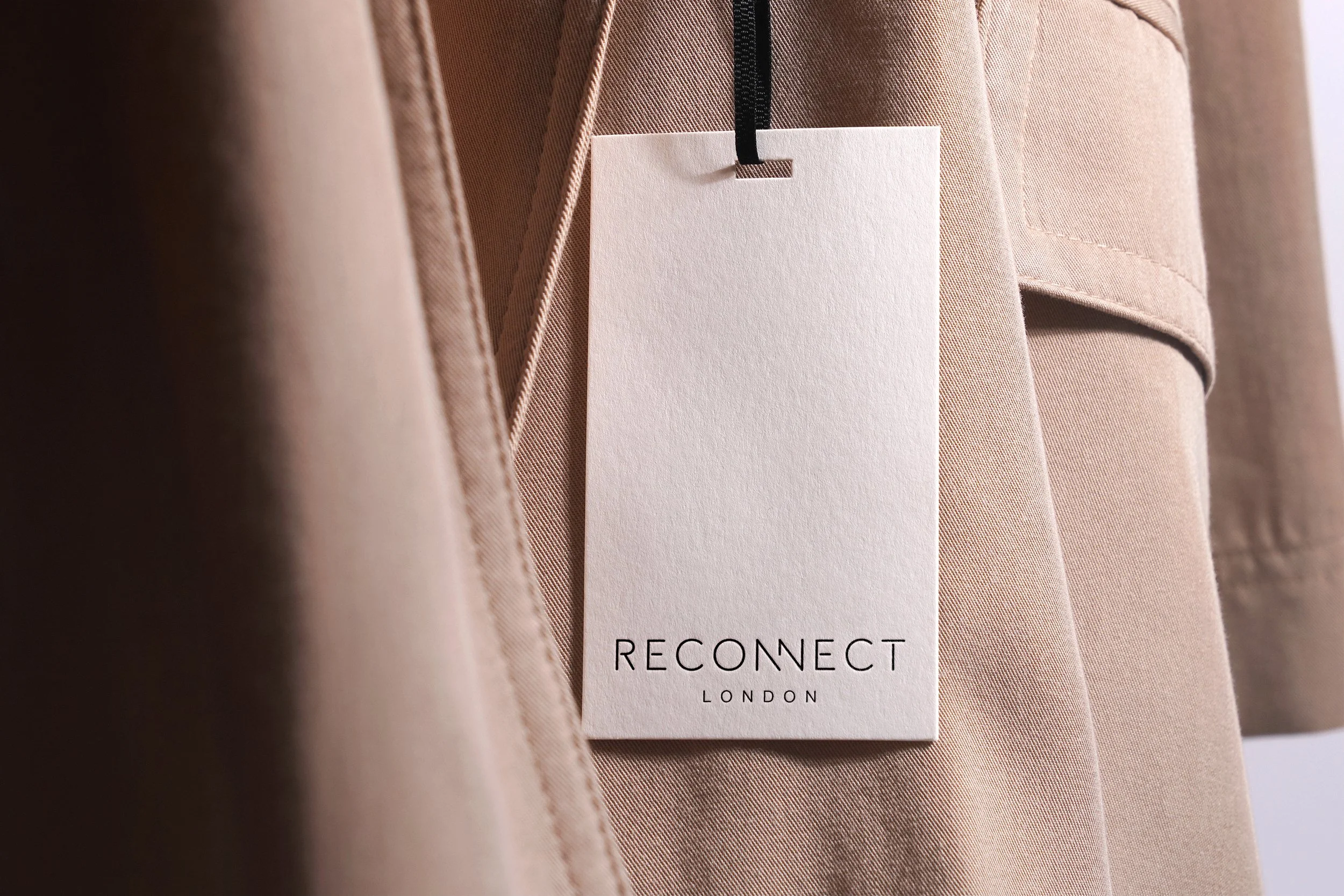 A beige coat with a white tag attached that reads 'RECONNECT LONDON'.