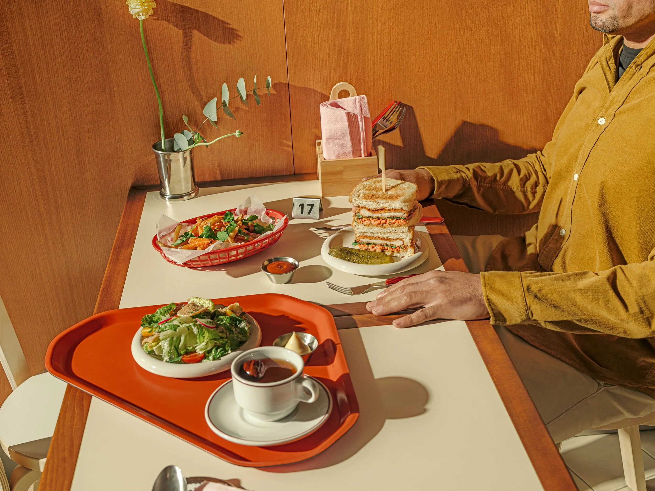 A man in a mustard-colored shirt sitting at a restaurant table with various dishes including a salad, a sandwich, a cup of coffee, and a bowl of soup, with a small vase holding a flower in the background.