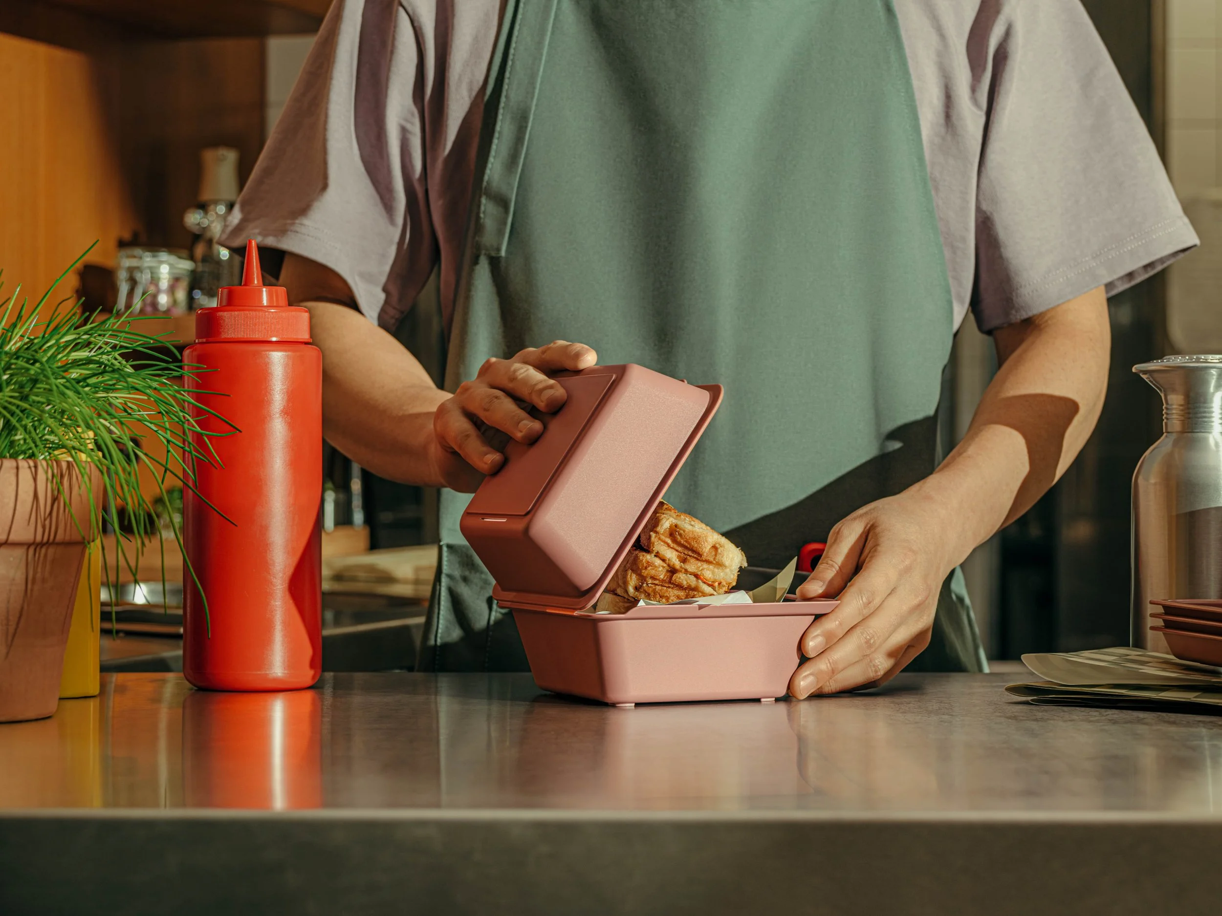 Person in a gray shirt and green apron opening a pink bento box with fried food inside on a kitchen counter.